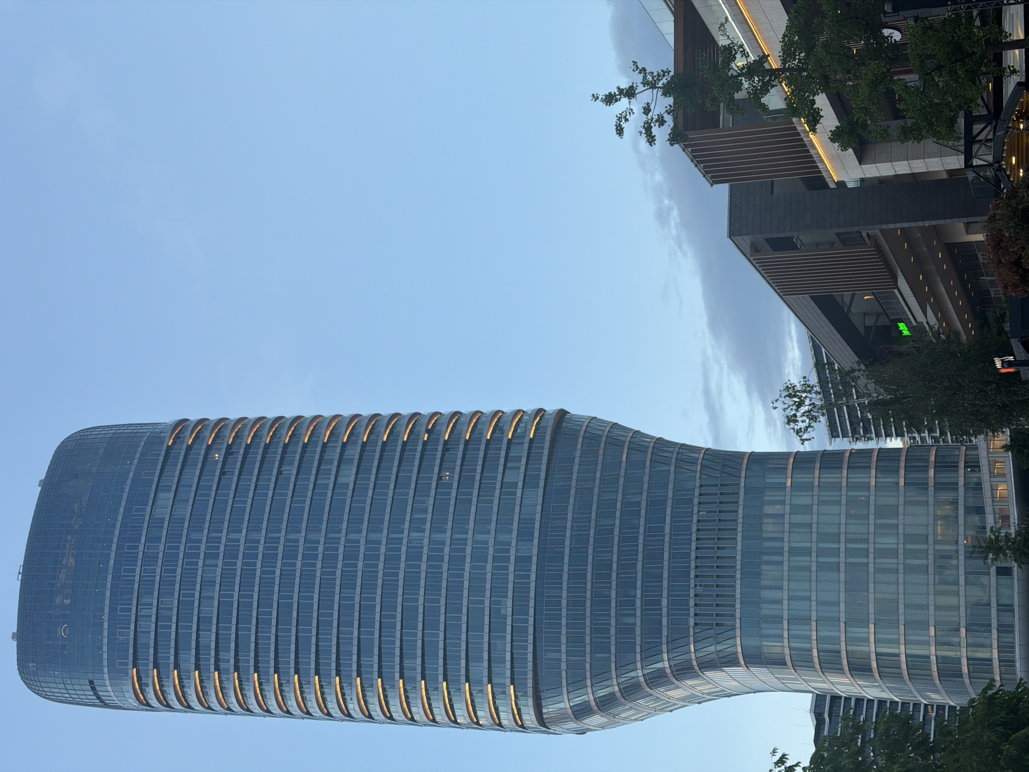 The image shows a modern, high-rise building in Belgrade, Serbia. The building features a unique, hourglass-shaped design with a glass facade and horizontal bands with golden light. The structure appears to be located in an urban setting, as there are trees and parts of other buildings visible around it. The sky is light blue.