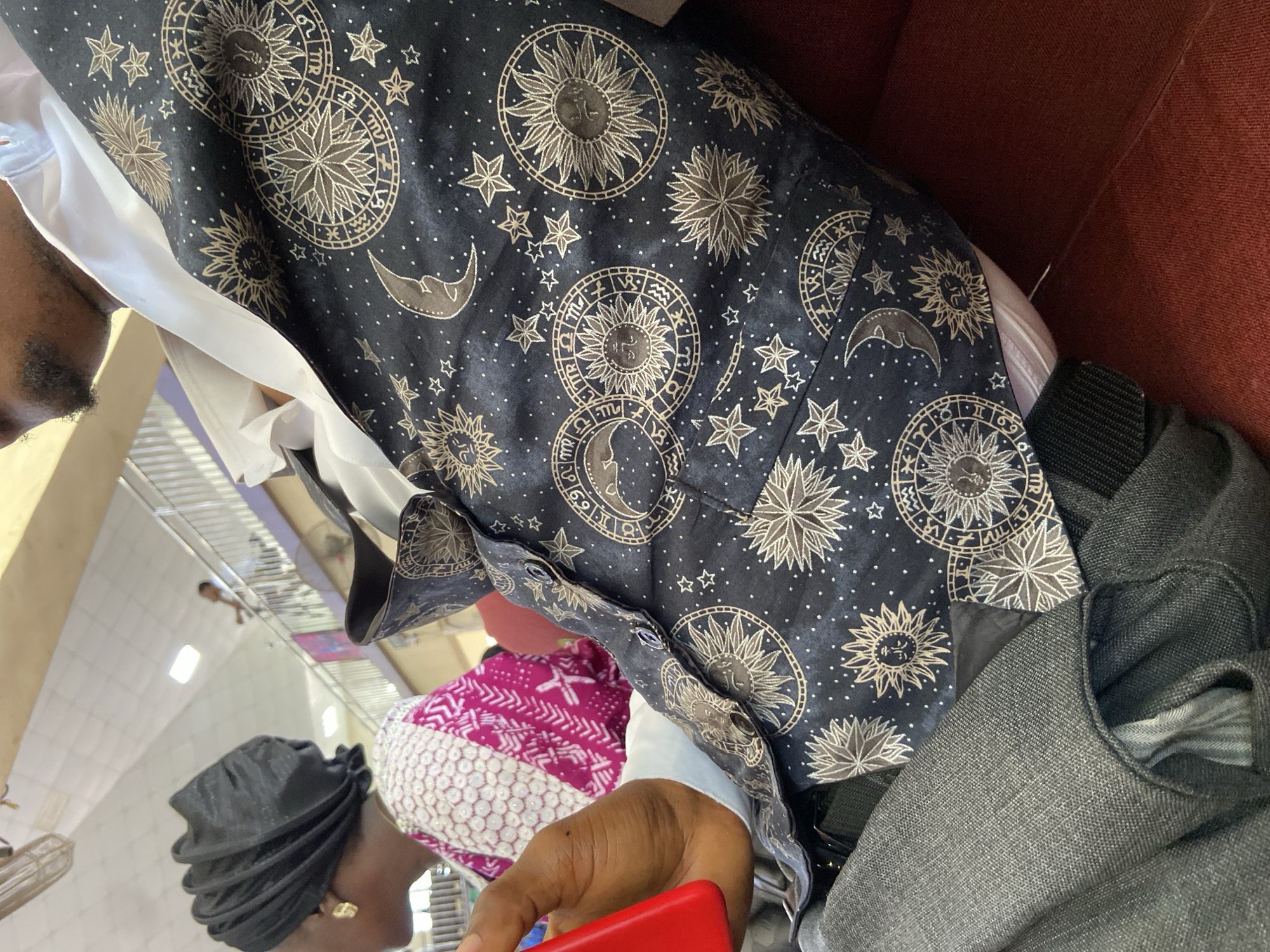 The image shows a person wearing a black shirt with celestial designs (suns, moons, stars, and zodiac signs). They are seated, with a dark grey denim or similar material bag next to them. Another person with a black head covering and pink/white patterned fabric around their head is partially visible. The setting appears to be indoors, possibly a waiting area in a location such as Mararaba, Nigeria, evidenced by the flooring pattern. Another person is walking in the distance along a shiny tiled floor. A red object is held in the foreground.