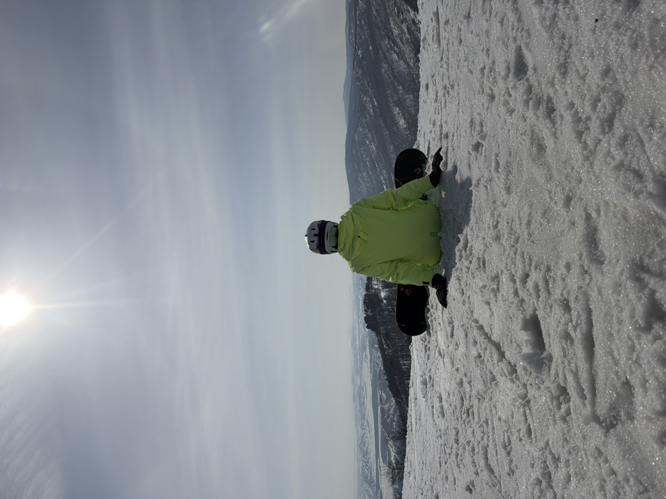 An individual is seated on a snow-covered slope in Zelenaya Polyana, Russia, facing away from the camera and towards a distant mountainous landscape. The subject wears a neon yellow winter jacket, black gloves, and a white helmet with a dark band. A black snowboard, displaying a subtle graphic on its tail, is propped against the individual's back. The foreground consists of granular snow. The background reveals a vast panorama of snow-dusted hills and mountains, many covered in dark trees, under a pale blue sky. A bright sun, positioned centrally in the upper frame, generates noticeable lens flare.