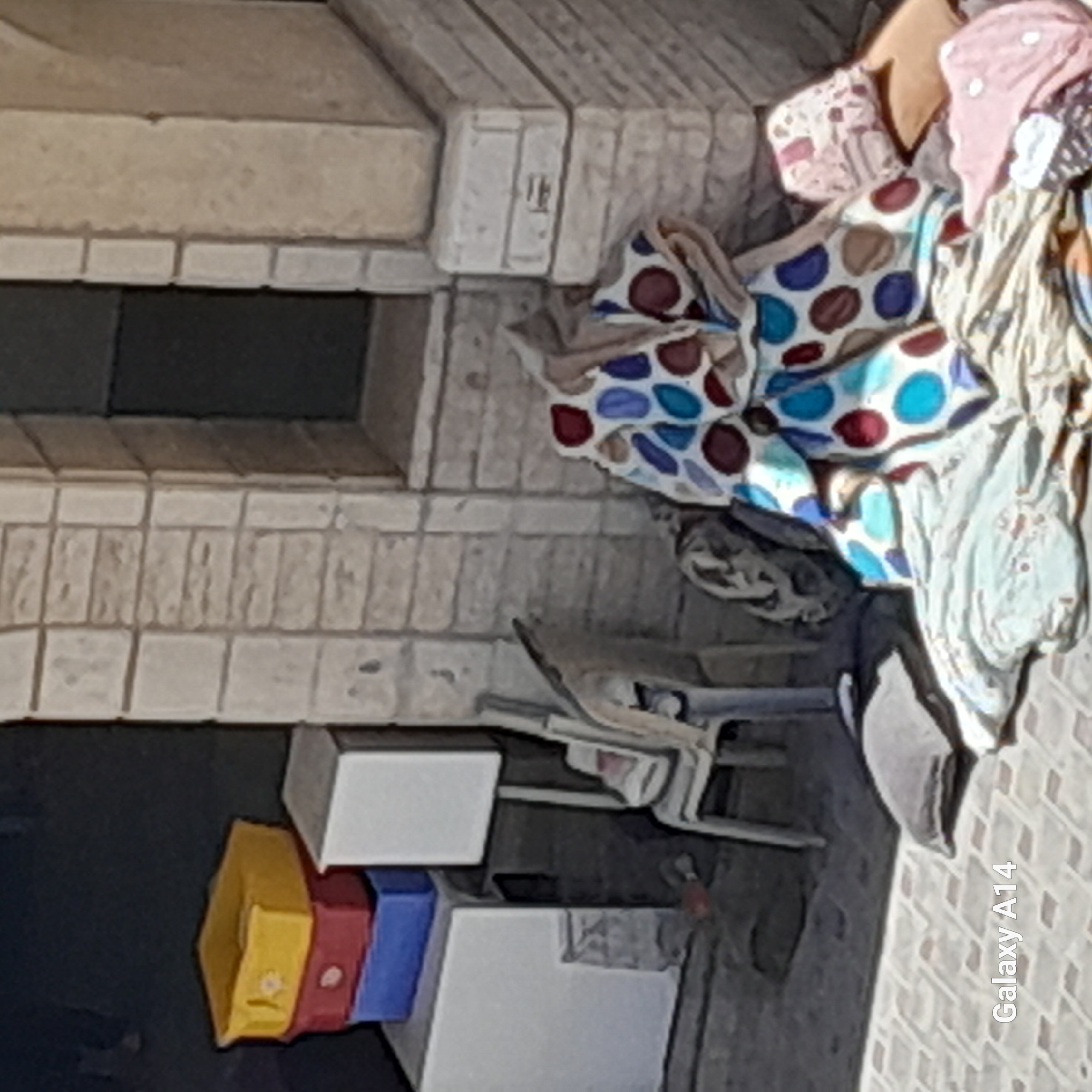 The image depicts an outdoor scene in Khan Yunis, Palestinian Territories, with a focus on a collection of household items and storage containers. In the background, a building constructed of light-colored bricks with a dark doorway is visible. In the foreground, a white plastic chair is positioned near a stack of colorful plastic containers, including yellow, red, and blue ones. To the right of the chair, a pile of blankets and linens, some with a polka dot pattern in shades of blue, red, and brown, are spread out on the ground. A white box or storage unit is also visible, as are two pillows on the pavement. The ground is paved with square tiles. The lighting suggests it is daytime.