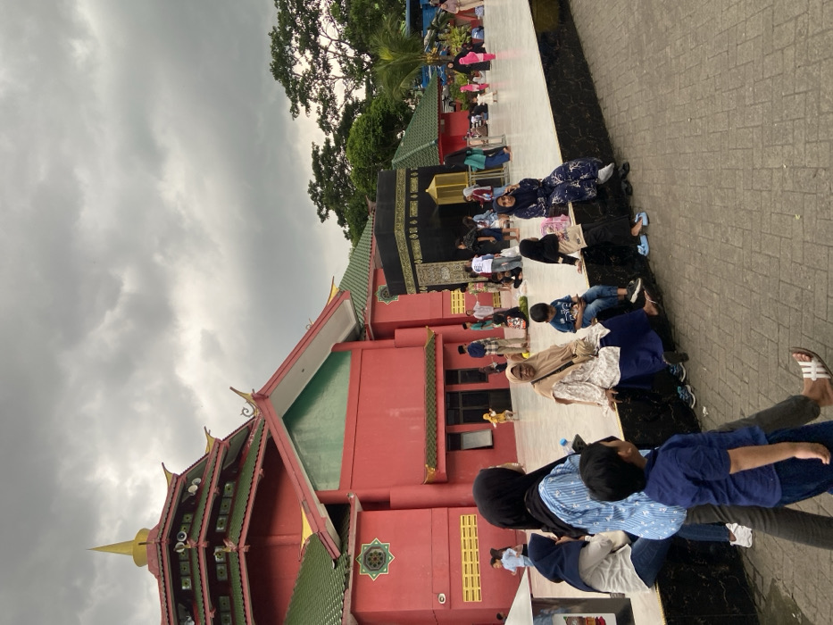 The image depicts an outdoor scene in Pandaan, Indonesia, under an overcast sky. A prominent multi-tiered red building with green and gold roofs, featuring traditional pagoda-like architecture and decorative elements, occupies the left and central background. To the right of this building, a large, black, cube-shaped structure with gold calligraphic patterns, resembling the Kaaba, stands on a light-colored paved area.
Numerous individuals, including adults and children, populate the scene. In the immediate foreground, a person's legs and feet, wearing white flip-flops, are visible. Moving backward, a woman wearing a blue patterned shirt and black hijab walks towards the right, holding a plastic bottle, alongside a young boy in a blue t-shirt and dark pants. To their left, another woman in a blue hijab and white top sits on a dark-colored raised ledge.
Further along this ledge, to the right, a woman in a beige hijab and patterned white top with a blue skirt sits, looking towards the viewer. A young boy in a blue t-shirt and jeans sits next to her, looking down at a mobile phone. Adjacent to them, another woman in a dark blue patterned dress and hijab sits, facing right. A young girl in a black top and jeans sits near her.
The large paved area in front of the red building and surrounding the Kaaba replica is populated by a crowd of people. Individuals are observed walking, standing, or sitting. Several children are present, some appearing to be playing. Trees and other vegetation are visible in the far background on the right side of the frame.