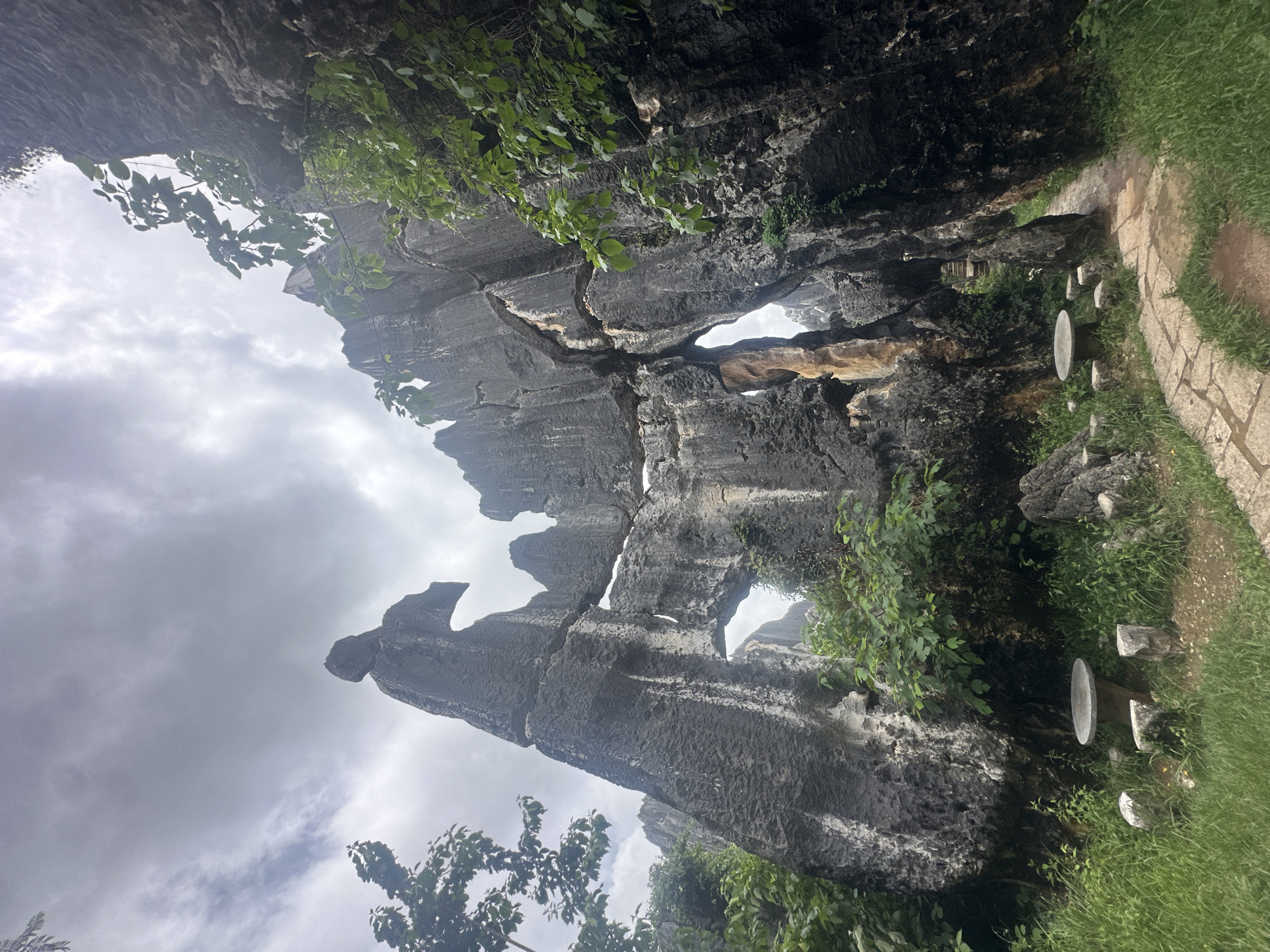 The image captures a natural rock formation in the Stone Forest, a karst landscape in Yunnan Province, China, near the city of Kunming. The foreground features a stone pathway winding through the towering, jagged grey limestone pillars. To the left, a round stone table is accompanied by three stone stools, nestled amongst lush green grass and foliage. More stone formations rise majestically in the background, some with dramatic openings and sharp, eroded edges. Green leaves and branches from trees and plants are visible, some even growing on or near the rock formations, suggesting a humid, verdant environment. The sky above is overcast with grey clouds, casting a subdued light on the scene. There are no people visible in the image.