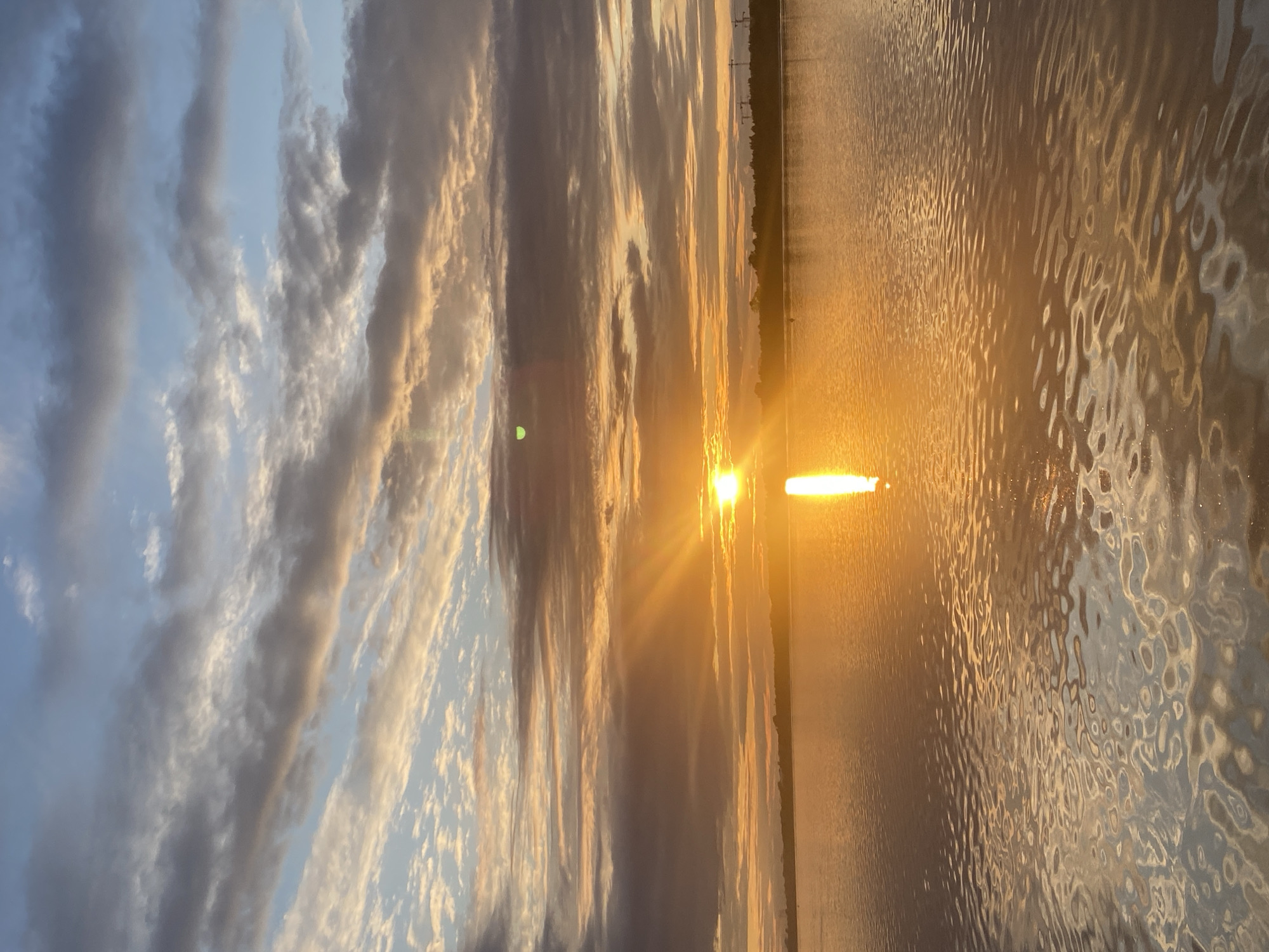 The image depicts a serene sunset over a body of water, likely the Kama River, near the city of Perm, Russia. The sky is a canvas of blues, grays, and oranges, with wispy clouds streaked across it, illuminated by the setting sun. The sun itself is a bright, golden orb, casting a radiant path of light across the calm water. The reflection of the sun shimmers on the rippling surface, creating a dynamic interplay of light and shadow. In the distance, a dark tree line marks the horizon, with a few indistinct structures, possibly industrial cranes, visible against the fading light. The overall atmosphere is one of tranquility and natural beauty.