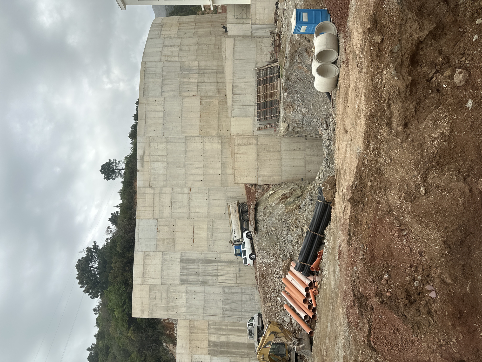 The image depicts a construction site in Budva, Montenegro. A large, recently constructed concrete retaining wall dominates the scene. The wall is segmented into rectangular panels. At the base of the wall, there are construction vehicles, including a dump truck, a pick-up truck, and an excavator. Stacks of black and orange plastic pipes are near the excavator. Further up, next to the wall are, a blue portable toilet, and three concrete pipes. The surrounding environment consists of a dirt slope, exposed rock, and trees on the hillside. The sky is overcast.