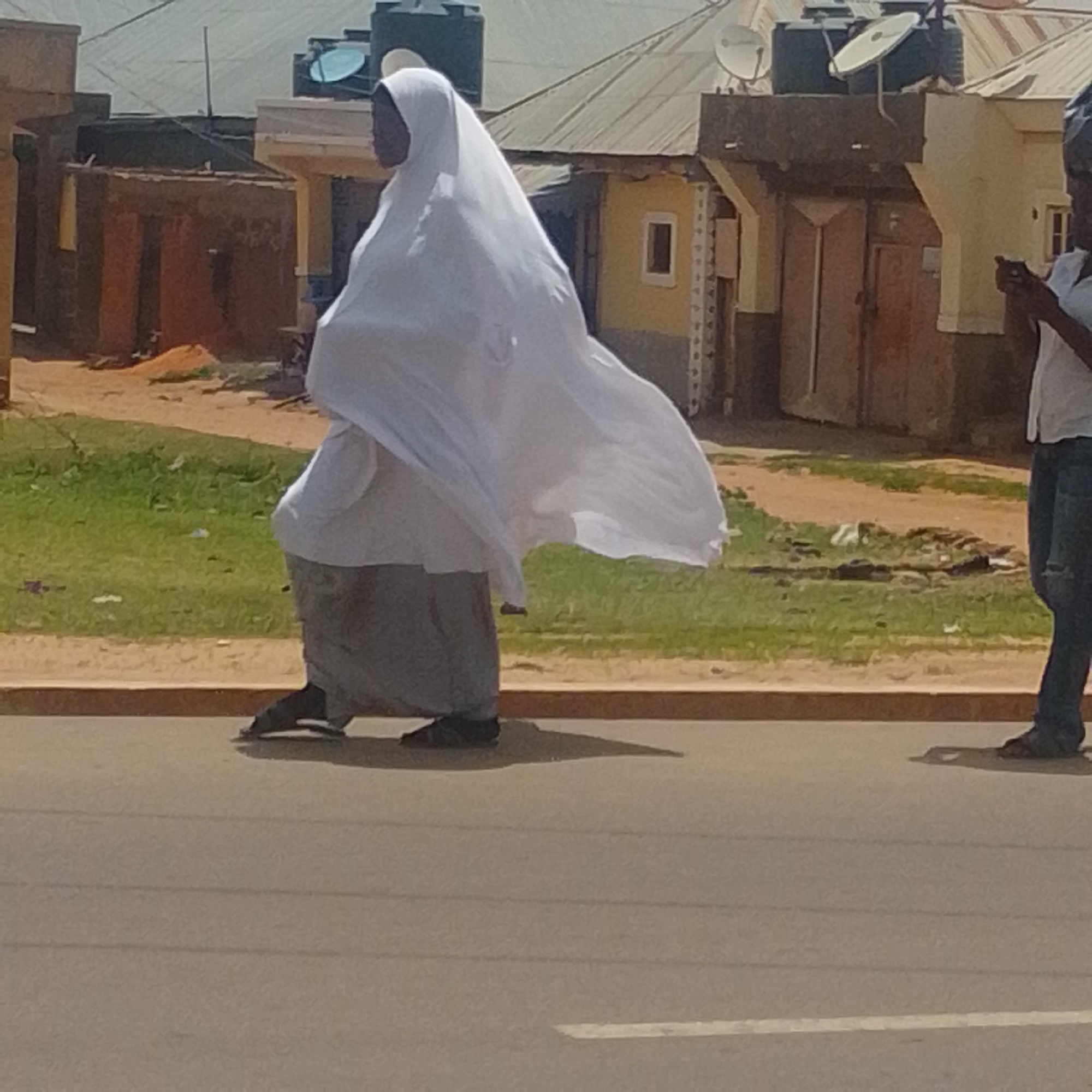 In Gombe, Nigeria, a person walks along a paved road in front of buildings and a grassy area. The individual is wearing a white headscarf and veil, along with a gray skirt. They are also wearing dark shoes. Their posture suggests they are walking forward, with their left leg extended.
Behind the person, there are several buildings with corrugated metal roofs. Some of the buildings are painted yellow, while others are made of reddish-brown materials. Water tanks and satellite dishes are visible on some of the rooftops, indicating the presence of modern amenities. The landscape appears somewhat arid, with patches of dry earth and sparse vegetation.
The time of day appears to be daytime, as there is ample natural light. The weather seems to be clear, with no visible clouds. The overall impression is one of everyday life in a Nigerian town, with a mix of traditional and modern elements. The person's face is not clearly visible, so their emotions cannot be determined. There is no visible text in the image.