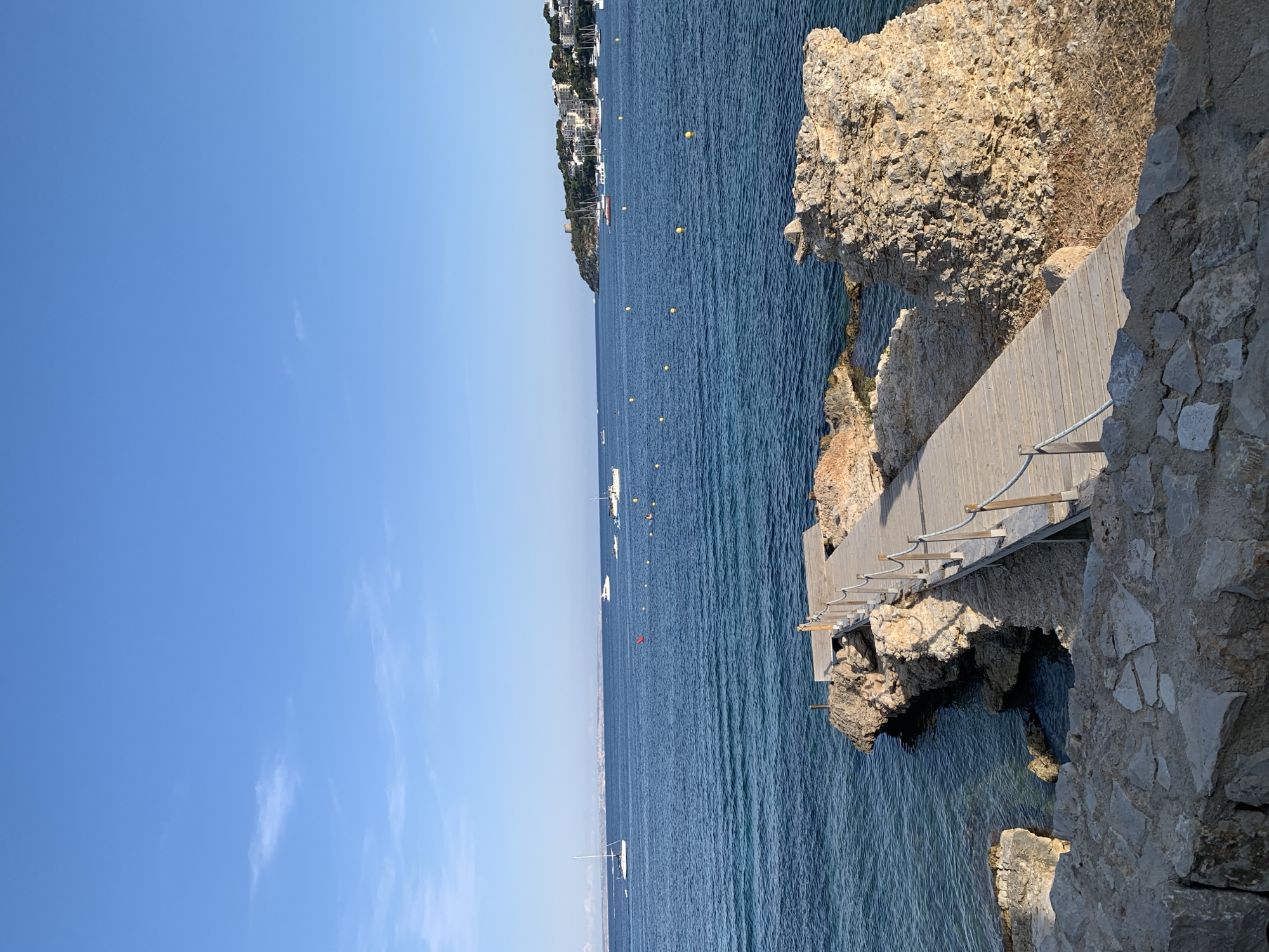 This media file captures a scenic view of a coastline with a wooden walkway extending over the water. The scene is set by the deep blue sea and a clear sky, suggesting a bright, sunny day, likely mid-day given the strong sunlight. The environment appears to be a coastal resort or a popular scenic spot, with buildings visible on a distant hillside, hinting at a Mediterranean or similar climate.
The main subjects are the natural elements: the rough, rocky cliff face on the right, the calm, blue sea, and the wooden walkway that seems to be a constructed path for visitors to enjoy the view or access the water. There are several yellow buoys dotting the water, possibly marking a swimming area or a navigational channel. A few sailboats and smaller vessels are scattered across the sea, indicating leisure activities.
The walkway is a notable detail, made of wooden planks and supported by simple structures, with a rope railing for safety. The stone wall along the right edge of the walkway adds to the rustic, natural feel of the location. The time of day is likely daytime, as evidenced by the clear blue sky and the bright lighting on the rocks and water. The overall mood is peaceful and serene, inviting viewers to imagine a relaxing day by the sea. No visible text is present in the image. The location context places this scene in Calvià, Spain, a region known for its beautiful coastlines and tourist attractions.