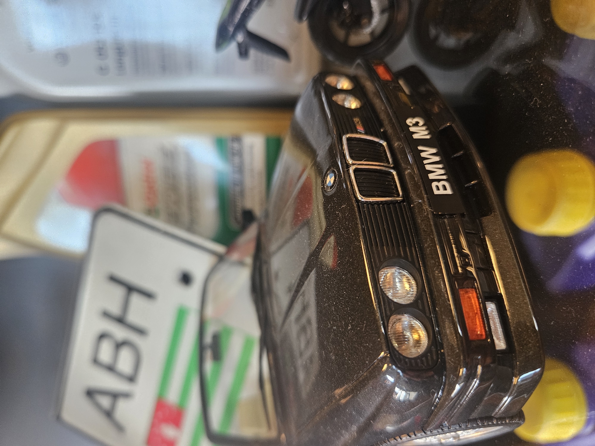 This is a close-up, eye-level, shallow depth of field shot of a black BMW M3 toy car. The car is dusty and positioned on a dark surface with two out-of-focus yellow caps in the foreground. Behind the car, partially visible, are several bottles, including one with a prominent red circular label and green stripes, and another clear bottle with text. A white sign with the letters "ABH" in black lettering and green and red stripes is also visible in the background, as is a blurred motorcycle. The scene appears to be indoors, with the lighting suggesting a controlled environment, possibly a display or collection. There are no people present. The location in Bemmel, Netherlands, is not discernible from the image itself.