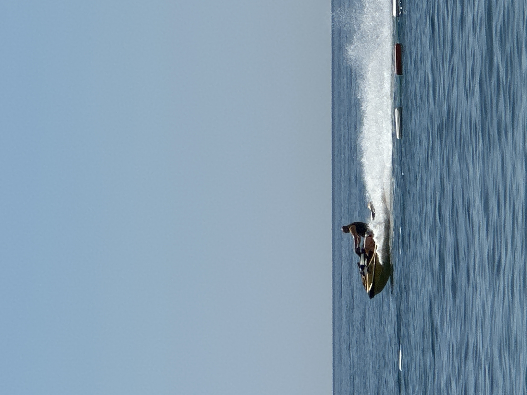 A man is riding a jet ski on a clear blue sea under a bright sky. The jet ski is yellow and black, and the man is wearing a dark t-shirt and shorts. He is creating a large spray of water behind him as he speeds across the water. In the distance, there are several buoys floating on the surface of the water, indicating a designated area or a race course. The sea is calm with gentle waves, and the horizon is visible as a clear line separating the sea from the sky. This scene is typical of recreational activities often found at coastal locations such as Boreti, Montenegro, known for its beaches and water sports.