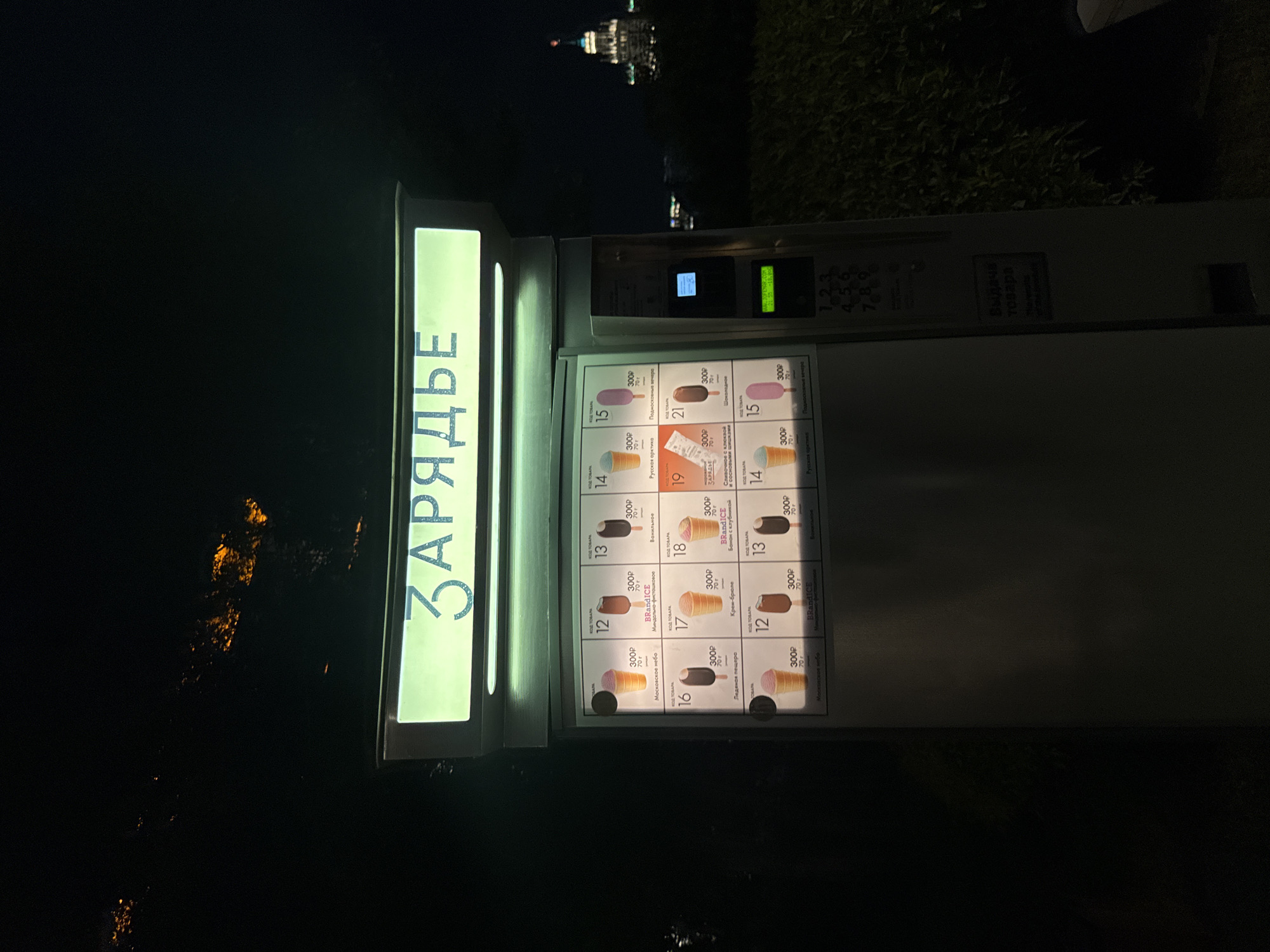 This is a nighttime, medium-shot, eye-level photograph of an ice cream vending machine located in Moscow, Russia. The machine has a lit-up sign at the top displaying the word "ЗАРЯДЬЕ" (ZARYADYE) in Cyrillic letters. Below the sign, a screen displays various ice cream options with their corresponding codes, prices (300₽ for 70g), and names. Examples of flavors include "Московское небо" (Moscow Sky), "Миндально-фисташковое" (Almond-Pistachio), "Ванильное" (Vanilla), "Русская практика" (Russian Practice), "Крем-брюле" (Crème brûlée), "Шоколадное" (Chocolate), and "Сливочное с клюквой и сосновыми шишками" (Cream with Cranberry and Pine Cones).
To the right of the ice cream display, there is a control panel with a small screen, a slot for payment, and a keypad with numbers 1 through 9. A label below the keypad reads "Выдача товара" (Goods Dispensing). In the background, out of focus, is a tall building with a spire topped by a red flag, likely a landmark in Moscow. The surrounding environment is dark, with some foliage visible to the right of the vending machine. The overall impression is of an urban setting at night, with the brightly lit vending machine being the focal point.