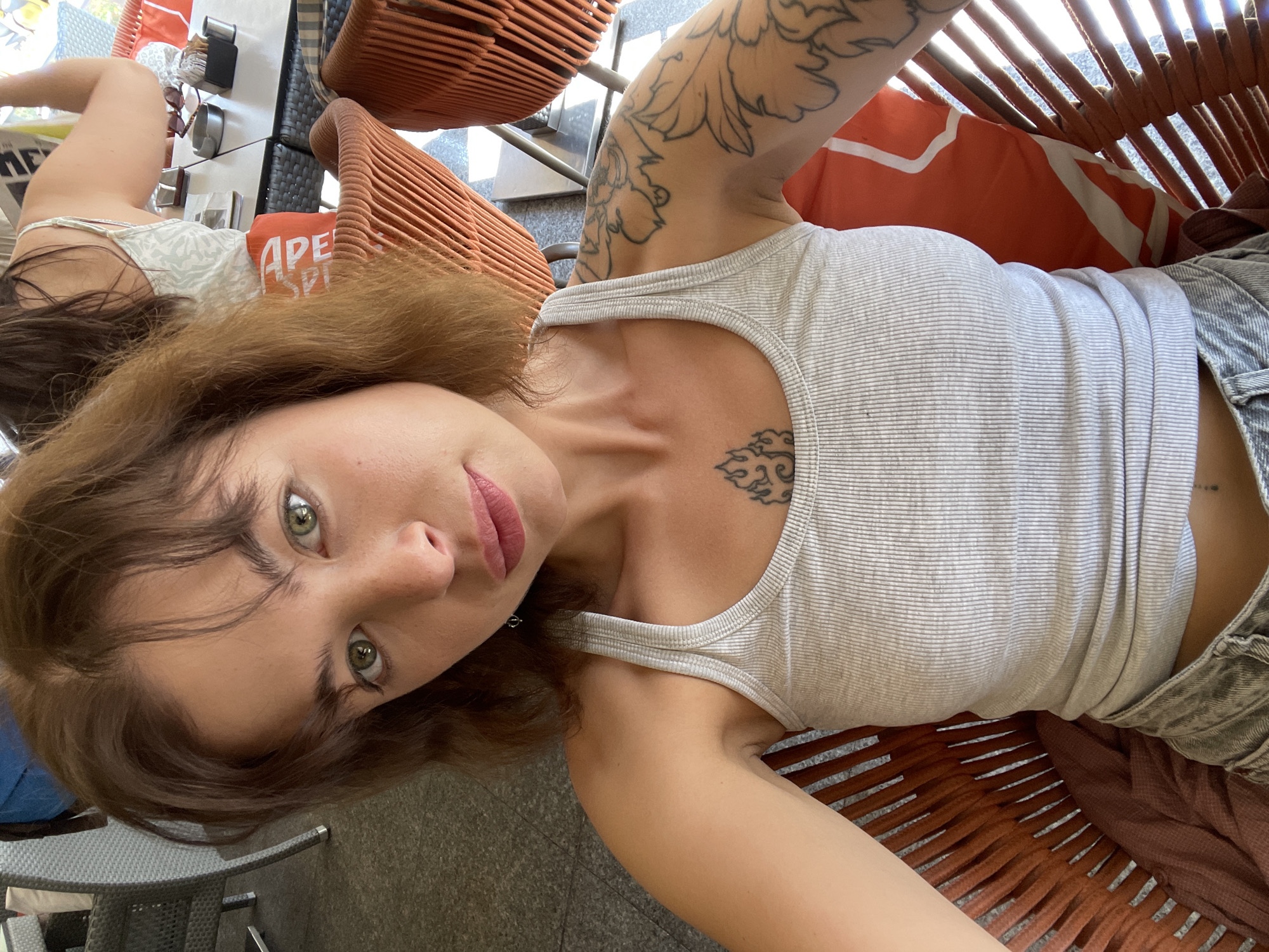 A young woman with light brown hair and green eyes is captured in a selfie taken outdoors, likely at a cafe in Budva, Montenegro. She is seated in an orange woven chair, with another person visible in the background. The woman is wearing a light grey ribbed tank top and denim shorts. She has tattoos on her upper chest and left arm. The background shows a table with what appears to be restaurant settings, and other outdoor seating. The overall scene suggests a casual, relaxed atmosphere, possibly during the daytime given the natural lighting.