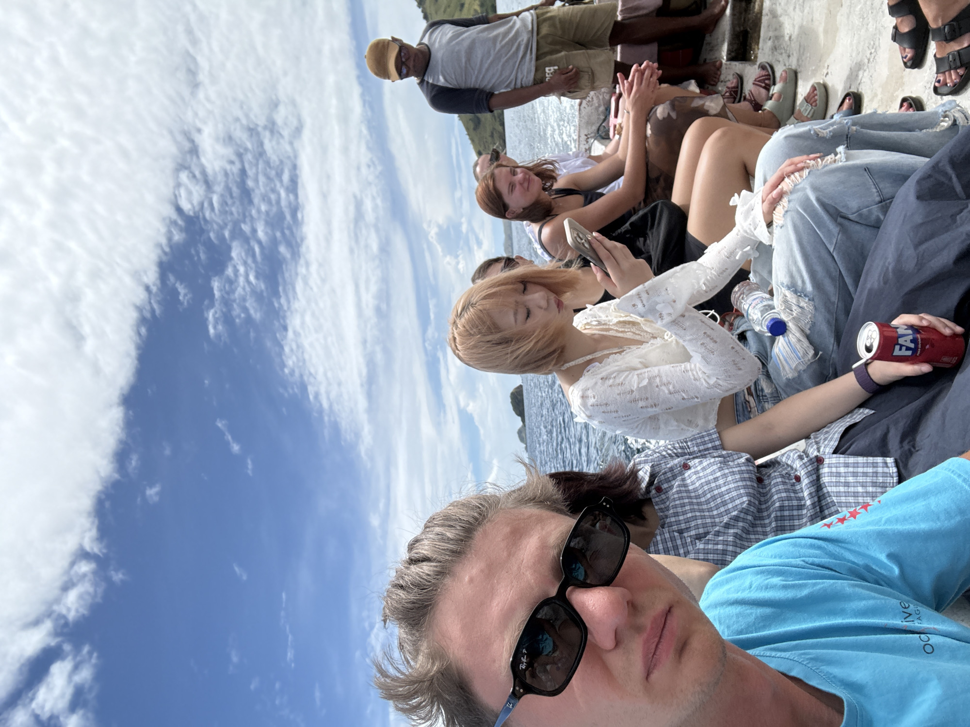 A medium, eye-level shot captures a group of individuals seemingly on a boat against a backdrop of water and cloudy sky. A man wearing sunglasses and a blue T-shirt is in the foreground. Behind him are other people, including one woman with blonde hair wearing a white lace top, another with auburn hair in a black tank top, and a man wearing a yellow cap, gray shirt, and tan shorts. The setting could potentially be in Unknown, Indonesia, based on water and landscape, but there are no identifying features. Several people are holding or looking at their phones. Other details in view include a water bottle, a can of soda, and assorted footwear.