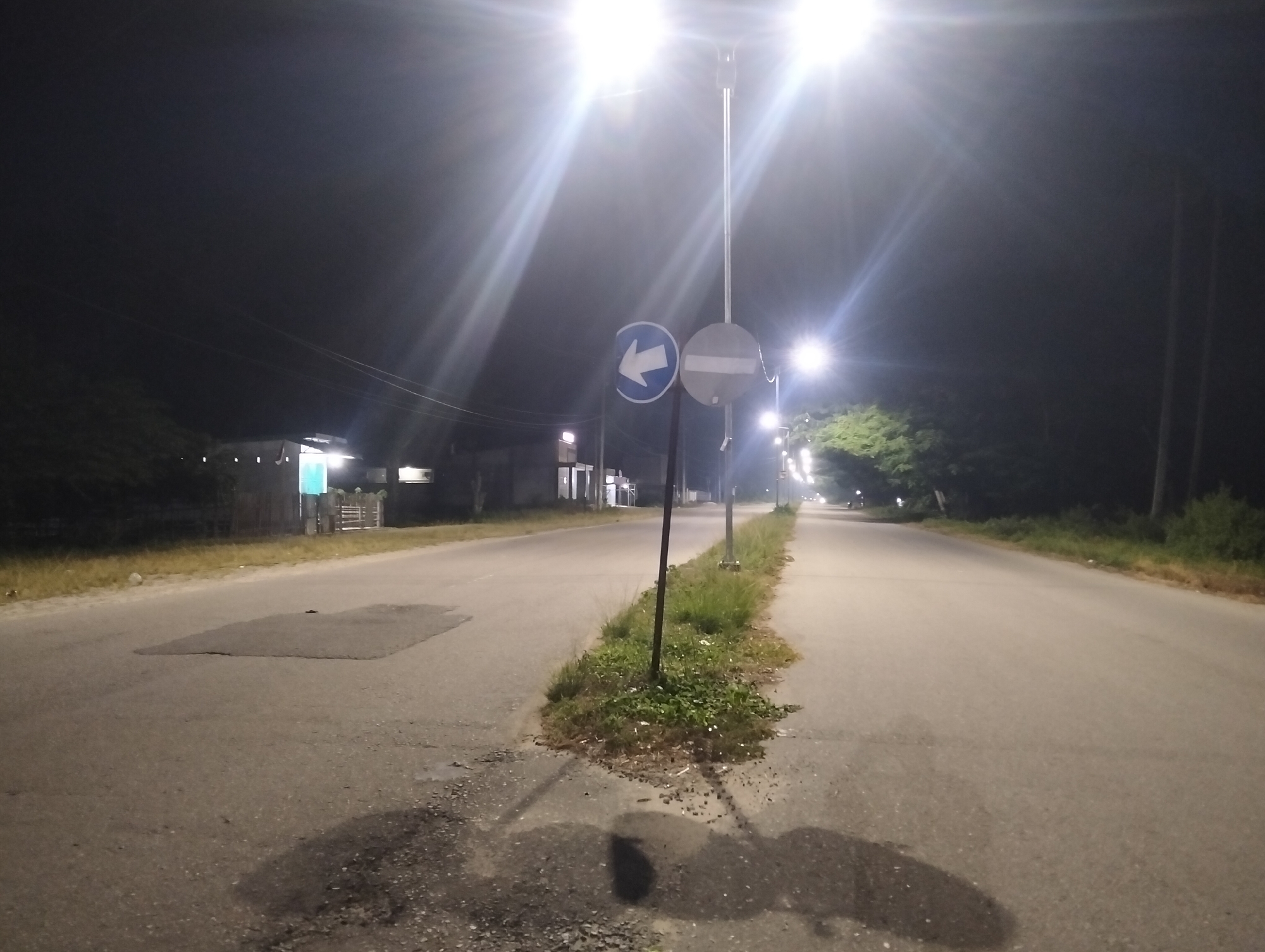 The image depicts a nighttime scene on a paved road in Kampal, Indonesia. The road is illuminated by streetlights, which create significant glare and lens flare, obscuring some details. On the left side of the road, there are buildings with illuminated windows and what appears to be a fence. A patch of dark asphalt is visible on the road surface.
In the center of the image, a traffic signpost stands with two signs. The upper sign is circular and white with a red horizontal bar, indicating no entry or a prohibition. The lower sign is also circular and blue with a white arrow pointing to the left, signifying a mandatory turn. A narrow strip of grass and vegetation runs along the median of the road, bordered by the two lanes.
On the right side of the road, more streetlights are visible, and dense vegetation lines the roadside. The road extends into the distance, with faint lights suggesting further development or structures. The overall environment is that of a semi-urban or rural road at night, characterized by artificial lighting and natural elements. There are no people or vehicles clearly visible in the frame.