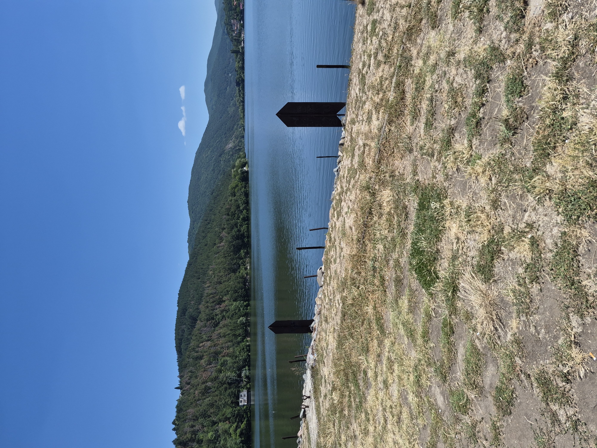 The image depicts a scenic view of Pancharevo Lake in Pancharevo, Bulgaria. The foreground is a dry, grassy embankment with scattered patches of dirt and sparse green vegetation. Several dark, upright posts, likely mooring posts, are visible along the shoreline. Two larger, decorative dark posts with intricate carvings stand prominently near the water's edge. Beyond the embankment, the calm, blue water of Pancharevo Lake stretches out, reflecting the clear blue sky. The opposite shore is lined with a dense, green forest that climbs the rolling hills. A white building is visible nestled amongst the trees on the left side of the lake, and more distant buildings and structures can be seen on the hills across the water. The sky is a vibrant, cloudless blue, with two small, wispy clouds appearing in the upper right portion of the frame. The overall scene is one of natural beauty and tranquility.