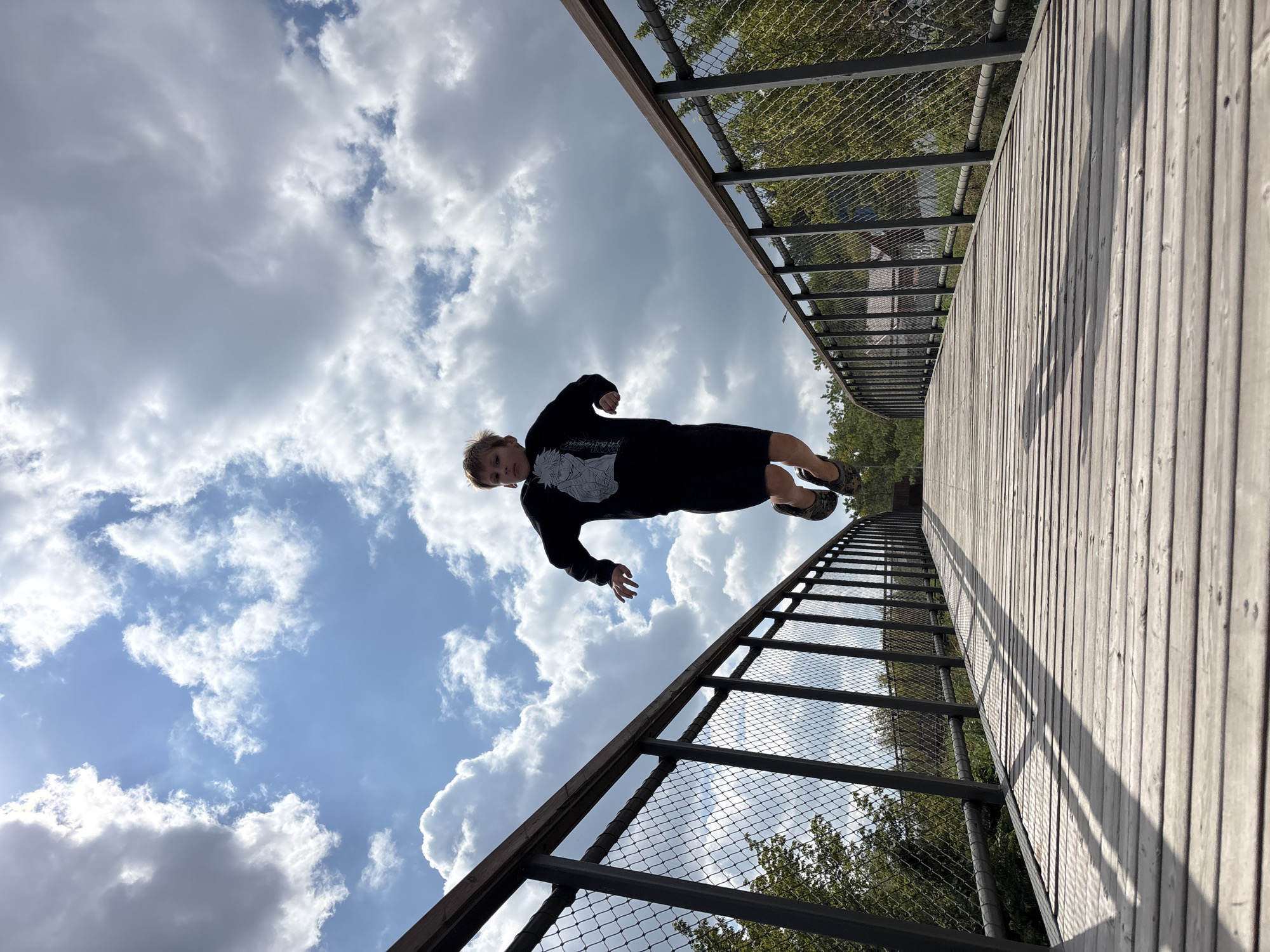 An outdoor, low-angle, eye-level, full shot shows a young boy mid-jump on a wooden footbridge under a cloudy sky. The boy, appearing to be between 8-10 years old, is wearing a black sweatshirt with a white graphic and black shorts. He is in the center of the frame, suspended in the air with his arms and legs extended. The bridge, made of light-colored wooden planks, extends away from the viewer and curves slightly to the right. Metal railings with a wire mesh infill line both sides of the bridge. Trees and some buildings are visible in the background beyond the railings. The sky is blue with scattered white clouds.