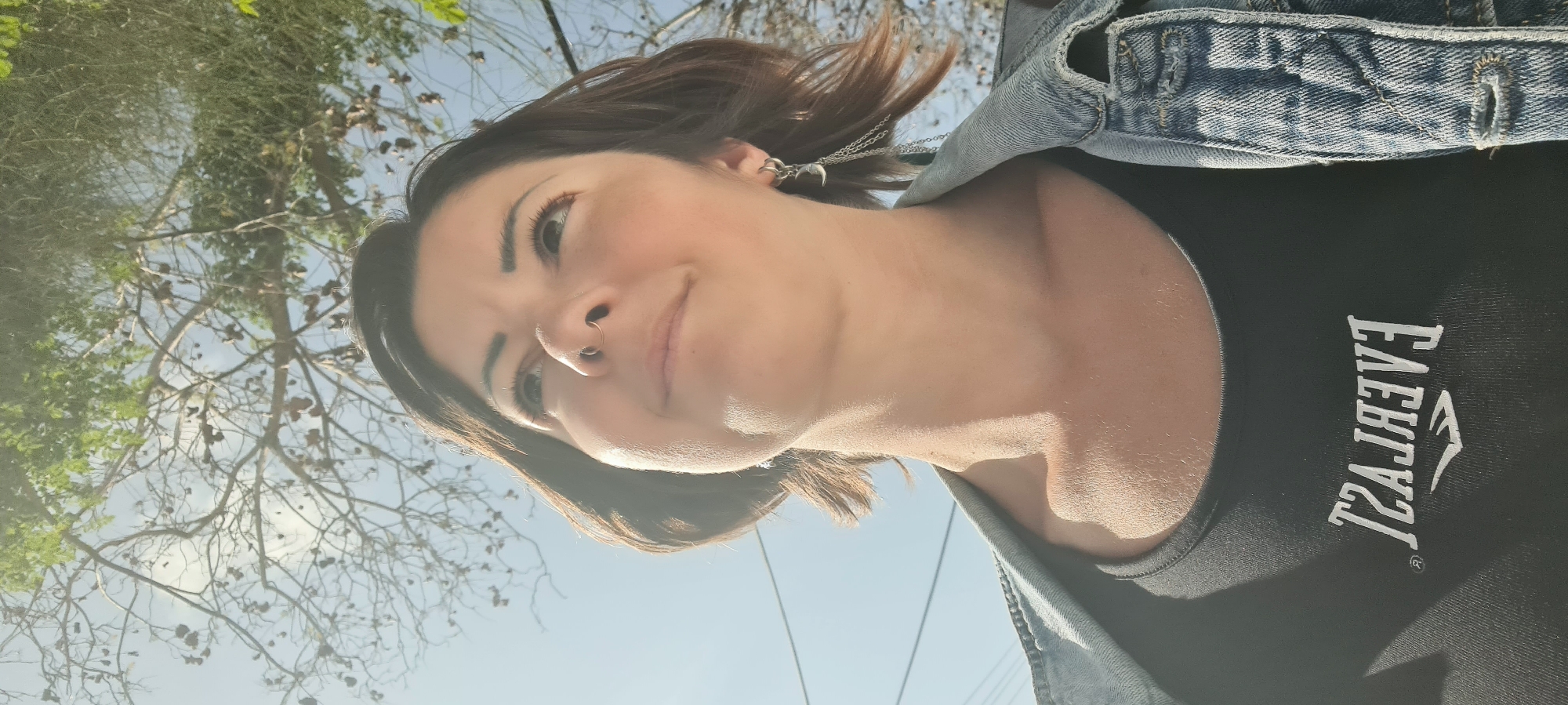 The image is a portrait of a woman, presented rotated 90 degrees counter-clockwise. When viewed upright, the woman is captured from the chest up, looking slightly upwards and to her right with a subtle, calm smile. She has medium-length dark brown hair, fair skin, a septum piercing, and a decorative earring featuring multiple small chains. She is wearing a dark (likely black) crew-neck top underneath a blue denim jacket. The word "EVERLAST" and its registered trademark symbol are visible in mirrored white text on her dark top.
The background consists of a clear, bright blue sky and trees. Some tree branches have green leaves, while others appear more sparse, showing bare branches with brown seed pods or dried foliage. The strong, direct sunlight illuminates her face and neck, indicating a sunny outdoor setting, likely during the midday or early afternoon. The overall scene suggests a casual moment captured in an open-air environment, consistent with the Lachish Regional Council, Israel.