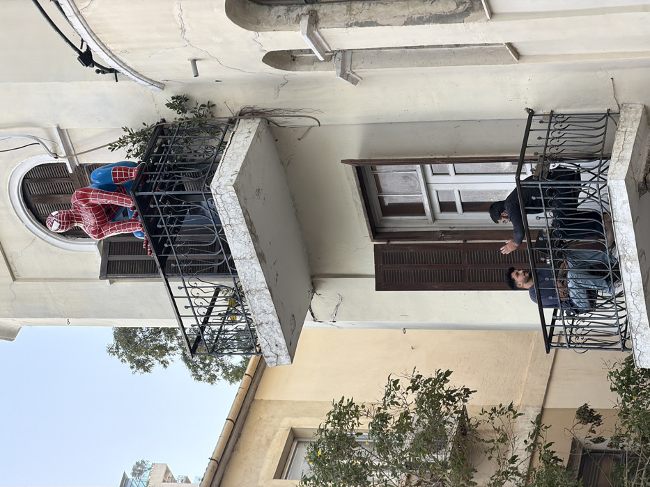 The image presents a section of an aged, light-colored residential building in Tel-Aviv, Israel, featuring two vertically stacked balconies with black wrought-iron railings.
On the upper balcony, a life-sized figure of Spider-Man, dressed in its characteristic red and blue suit, is crouched on the railing. The figure's hands are positioned on the railing, and its head is turned slightly to the left. The concrete surface of this balcony displays notable cracking and weathering. Behind the Spider-Man figure, a window with open dark-brown wooden shutters is visible, featuring an arched top.
On the lower balcony, two adult males are seated on chairs, facing each other and engaged in conversation. The male on the left has dark hair and a beard, wearing a dark long-sleeved shirt and blue jeans, and is looking at the other individual. The male on the right, also with dark hair and a beard, wears a dark long-sleeved garment and has his right hand raised with an open palm, indicating a gestural action during speech. Behind them, a window with open dark-brown wooden shutters reveals a white-framed window.
The building's cream-colored facade exhibits cracks and signs of deterioration. To the immediate left, a portion of an adjacent building, lighter yellow-beige in color, is partially visible, with green foliage in the foreground. The sky is clear and light, suggesting daytime.