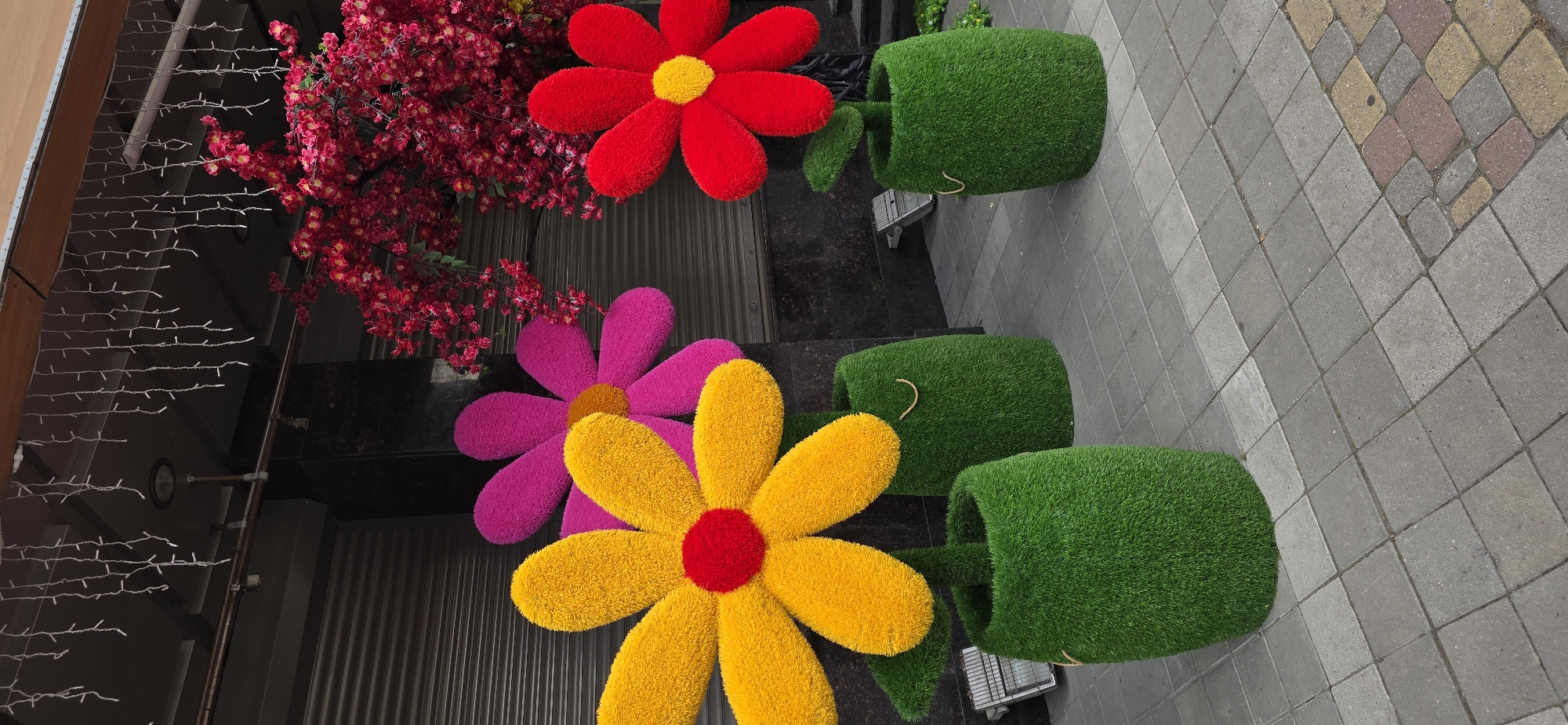 Three oversized, artificial flowers stand in green, grass-covered pots on a paved sidewalk. The flower on the left is yellow with a red center, the middle flower is pink with a yellow center, and the flower on the right is red with a yellow center. Behind the flowers, a building with closed gray shutters is visible, with a dark facade and a wooden overhang adorned with white string lights. To the right of the flowers, a dense arrangement of artificial red flowers on branches is suspended, creating a canopy effect. The sidewalk is paved with gray square tiles. The scene appears to be an outdoor decoration, possibly for a shop or event, in the city of Kislovodsk, Russia. No people are present in the image.