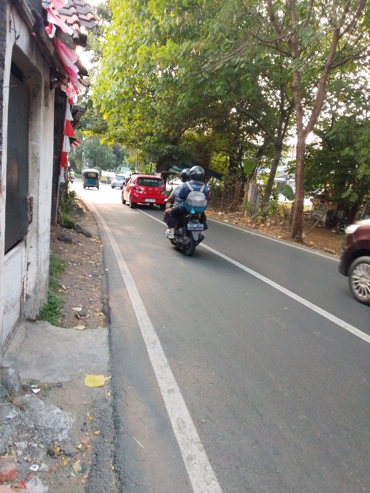 The image shows a street scene in East Jakarta, Indonesia. In the foreground, a motorcyclist wearing a helmet and a blue backpack is riding on the right side of the road. A portion of a brown car is visible on the far right of the image. Further down the road, a red car is parked on the left. To the left of the road, there is a building with a textured wall and a tiled roof, adorned with red and white decorations. Trees with green foliage line the right side of the road. The road surface is asphalt with a white line marking the lane. The overall scene appears to be an outdoor environment during daylight.
