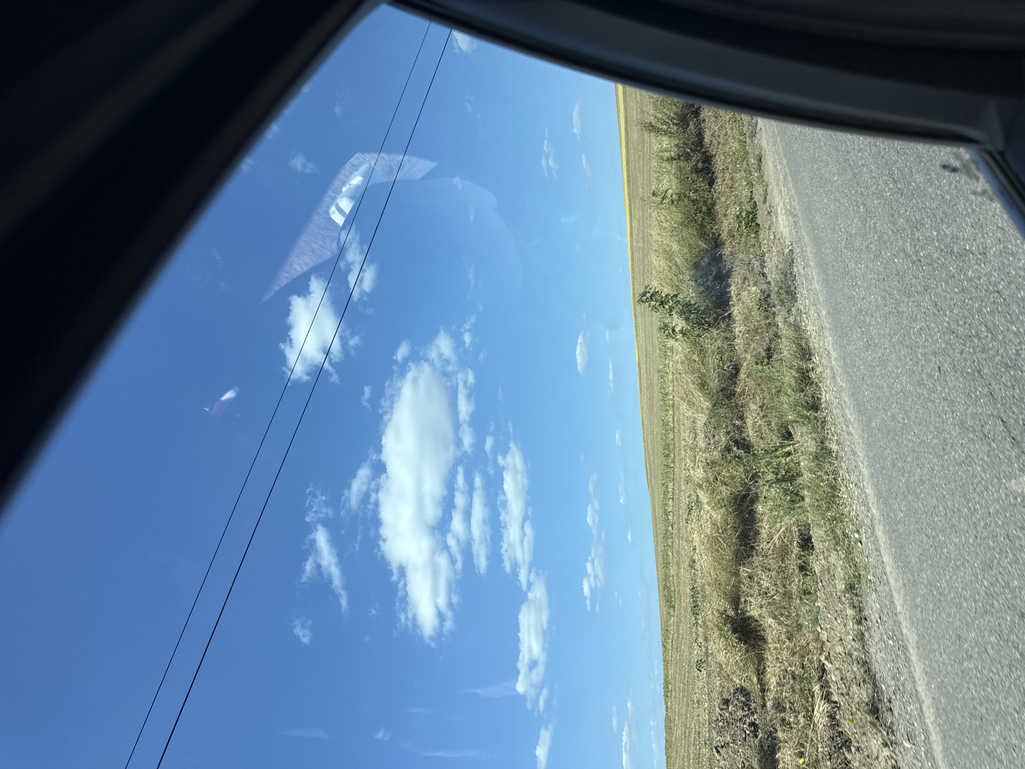 The image shows a view from a car window looking out at a rural landscape. The sky is bright blue with scattered white clouds. Two black electrical wires stretch horizontally across the upper portion of the sky. Below the sky, there is a field of dry, golden grass and some patches of darker soil. A small green bush is visible in the middle ground. A paved road runs along the bottom of the image, with the side of the car frame visible on the right. The scene appears to be in the city of Lehancea, Romania, given the context provided. There are no people or animals visible in the image.