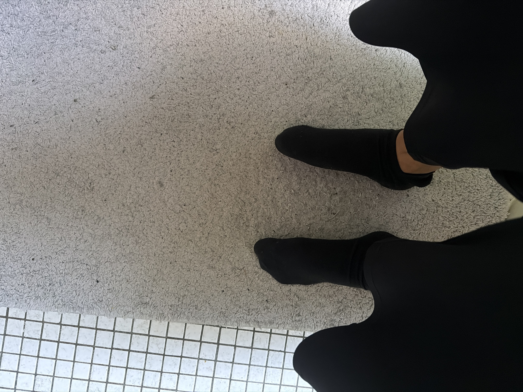 A first-person perspective shot shows two feet wearing black socks, positioned on a light grey textured rug. To the left of the rug, a section of white tiled flooring is visible, with a grid pattern formed by dark grey grout lines. The scene appears to be indoors, possibly in a bathroom or a room with a rug. There are no visible people other than the feet. The lighting suggests it is daytime. No specific landmarks or indicators for Nienburg/Weser, Germany are present in the image.