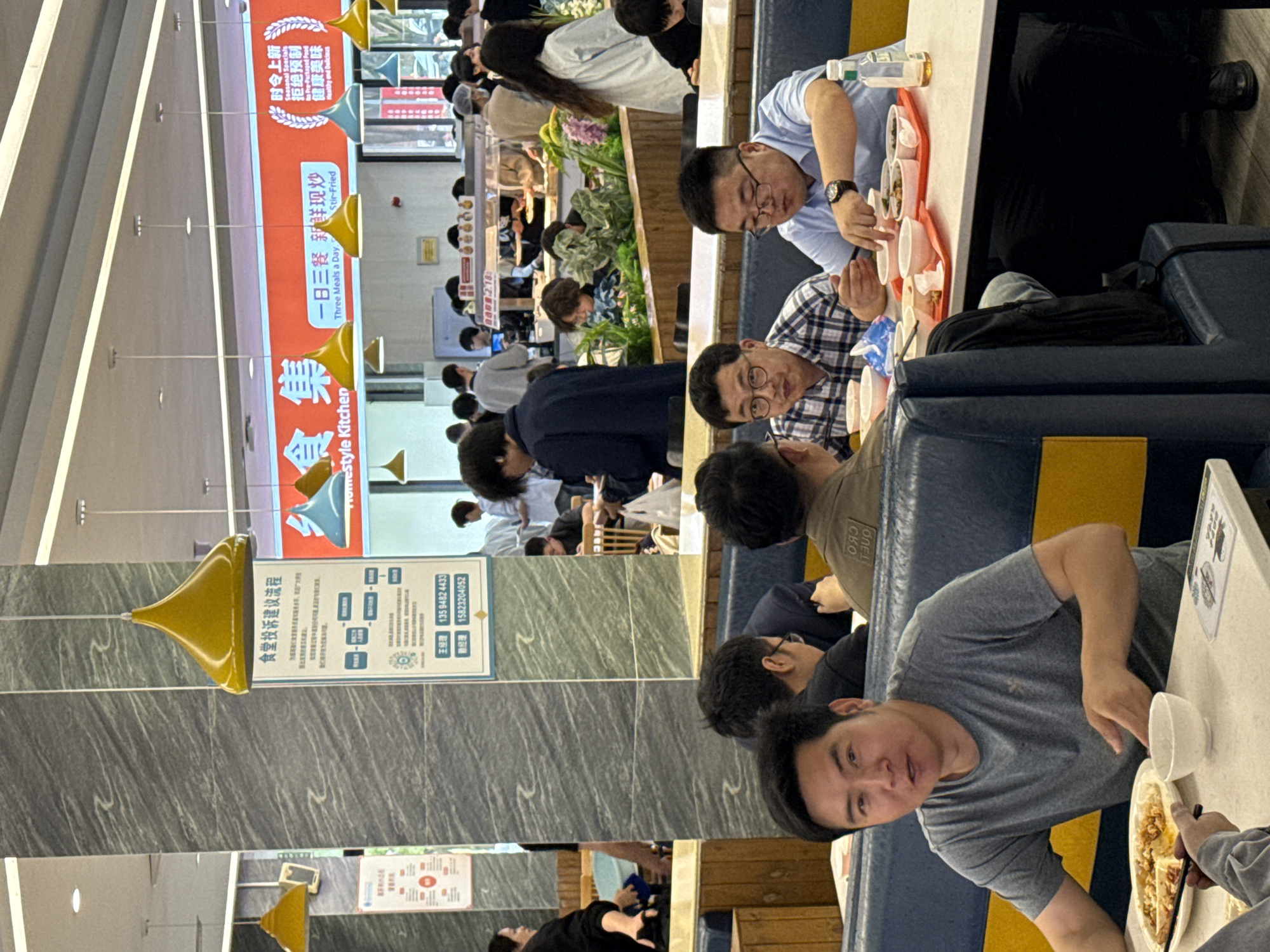 The image captures a bustling scene in a cafeteria, likely in a college or university setting based on the age of the individuals present. The location is Jiangjin, China, based on the Chinese text present in the signage. There are several groups of people sitting at tables, eating, and interacting with each other. Some individuals are wearing glasses. One person is wearing a light blue shirt and is looking at food with a plate of small bowls in front of them. Another person is wearing a blue and white plaid shirt, also focused on their food. There are several tables arranged with booth-style seating, a combination of blue and yellow. The environment includes signage with text in both English and Chinese, some of which promotes the cafeteria or its food options. One sign is related to restaurant complaints and contact information. The lighting is bright and the overall scene indicates a busy and active place for communal dining.