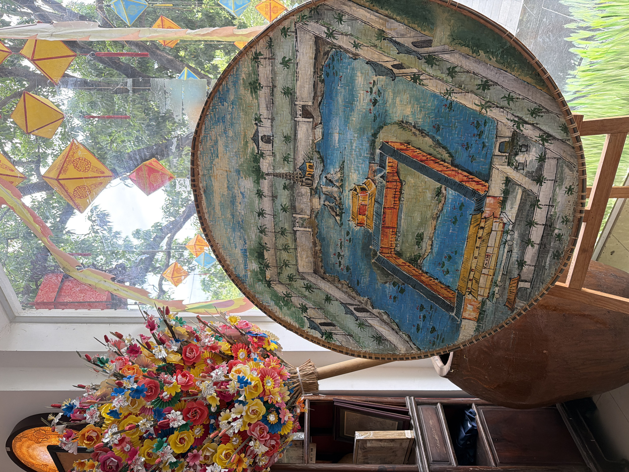 The image depicts an interior scene, possibly a shop or gallery, featuring a large, round, woven piece of art displayed on a wooden easel. The artwork itself is a painting on a woven surface, showing a bird's-eye view of a complex water system and buildings, possibly a historical or architectural site. The painting is detailed, with blue water, orange and yellow structures, grey walls, and green trees. To the left of the artwork, a tall wooden cabinet holds framed items, and a large, rustic, terracotta-colored pot stands next to it. In the foreground and to the left, a vibrant bouquet of artificial flowers in various bright colors, including red, yellow, pink, and blue, is arranged on a wooden pole. Behind the artwork and artificial flowers, a window reveals an outdoor scene with lush green trees and several colorful, square paper lanterns hanging, some yellow and some red, some adorned with red tassels. The lanterns suggest a festive atmosphere, possibly a celebration or market event. The light filtering through the window suggests it is daytime. The overall impression is one of cultural display and artistic presentation. The location is stated to be Thuận Hóa Ward, Vietnam.