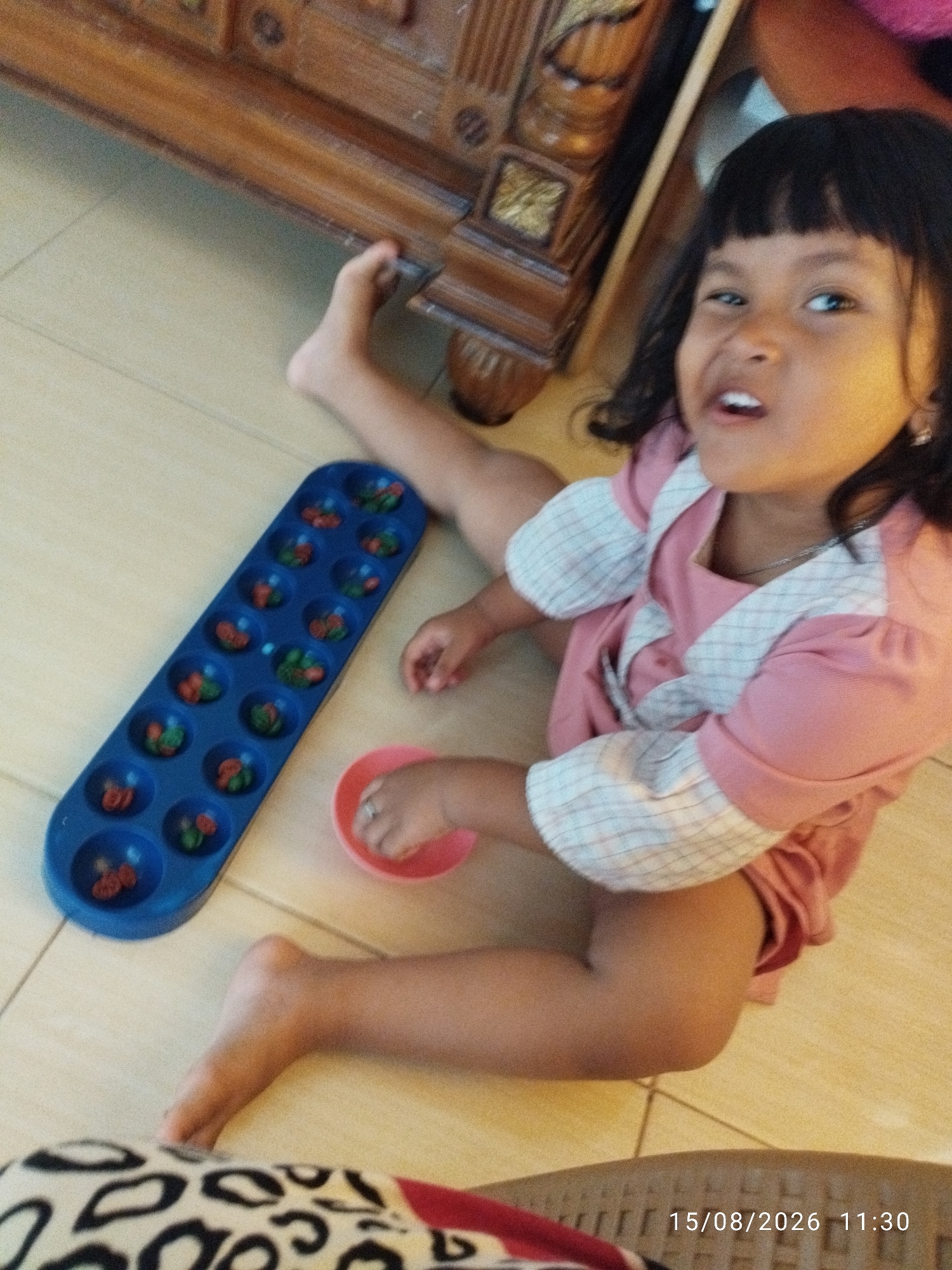 An overhead, eye-level shot shows a young girl with dark hair and bangs sitting on a tiled floor. She is wearing a pink dress with white plaid accents on the sleeves and collar. She is playing with a blue Mancala board, which is filled with red and green stones. Her right hand is near a small pink bowl, and her left foot is visible. In the background, a wooden piece of furniture is partially visible. The scene is indoors, likely in a home, as indicated by the flooring and furniture. A timestamp in the bottom right corner reads "15/08/2026 11:30". This event is presumed to have occurred in Bandar Lampung, Indonesia.