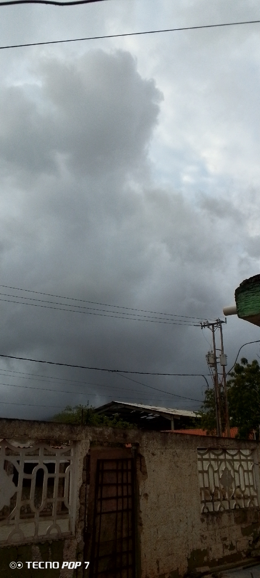 The image depicts a cloudy sky over a residential area in Altagracia, Venezuela. The sky is filled with dark, heavy clouds, suggesting an impending storm or overcast weather. Several power lines crisscross the frame, adding to the urban or suburban setting.
In the foreground, there is a weathered concrete wall with a decorative metal gate on the left and a metal-barred door on the right. The wall appears to be part of a property boundary. Behind the wall, glimpses of structures can be seen, including a building with a corrugated roof and some trees. A utility pole with electrical equipment is also visible, further indicating an inhabited area.
The time of day is indeterminate due to the overcast conditions, but the lighting suggests it is daytime. The mood of the image is somewhat somber, largely due to the dark clouds. There are no people visible in the frame.
At the bottom left corner of the image, there is a watermark that reads "TECNO POP 7".