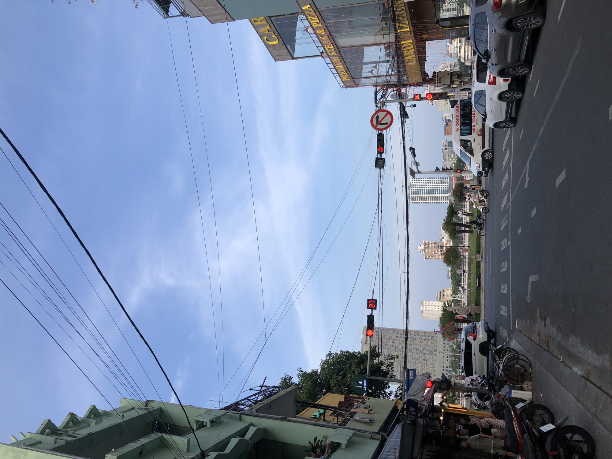 The image depicts a street scene in Đà Nẵng, Vietnam. A wide road with multiple lanes is visible, marked with white dashed lines and a pedestrian crossing. Several vehicles are present, including cars, a white van with "DOKKAEBI NHA TRANG" written on its side, and motorcycles parked along the sidewalk. A traffic light with a red signal is illuminated. On the right side of the street, a multi-story building features a sign that reads "CARDIPizza" and "PREMIUM STEAK & PIZZA." The building is decorated with red string lights. On the left side, a green building with a textured facade and balconies is partially visible. Power lines crisscross the sky, which is a clear blue with a few wisps of clouds. In the background, tall buildings and a bridge can be seen. The time of day appears to be late afternoon or early evening.