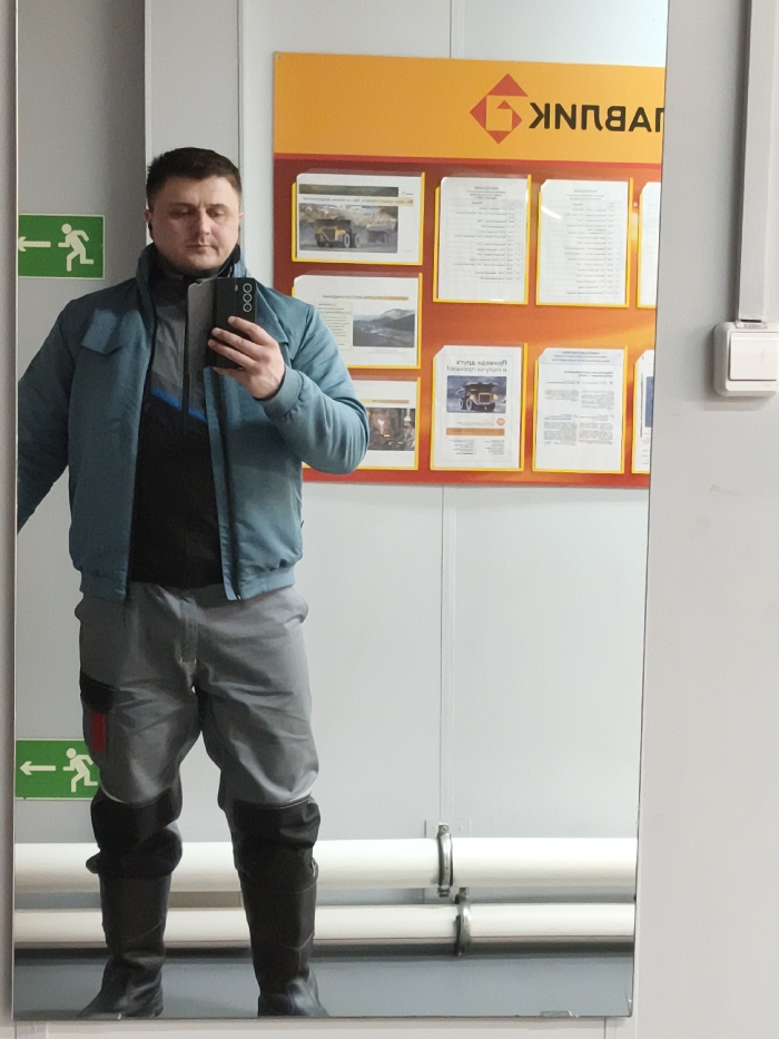 An adult male stands for a full-body mirror selfie indoors in the city of Im. Gastello, Russia. The subject, a Caucasian male with short brown hair, wears a teal-blue bomber jacket over a dark-colored zipped mid-layer with a blue stripe, grey work trousers featuring a black and red utility patch on the left thigh, and black tall boots. He holds a dark-colored smartphone with a light case in his right hand, angled towards his reflection. The background behind the subject's reflection consists of a white wall. Mounted on this wall is a large orange and red informational board displaying multiple documents in yellow-bordered clear plastic sleeves. Several visible documents feature images of large mining dump trucks in an industrial setting. A reversed Cyrillic logo, appearing as "KBNNPUH," is visible at the top right of the board. To the left of the reflection, two green "exit" signs with white running figures and left-pointing arrows are affixed to the wall. In the lower background, two horizontal white pipes secured by silver clamps are visible. A white electrical switch or outlet is mounted on a grey vertical panel to the right of the informational board's reflection.