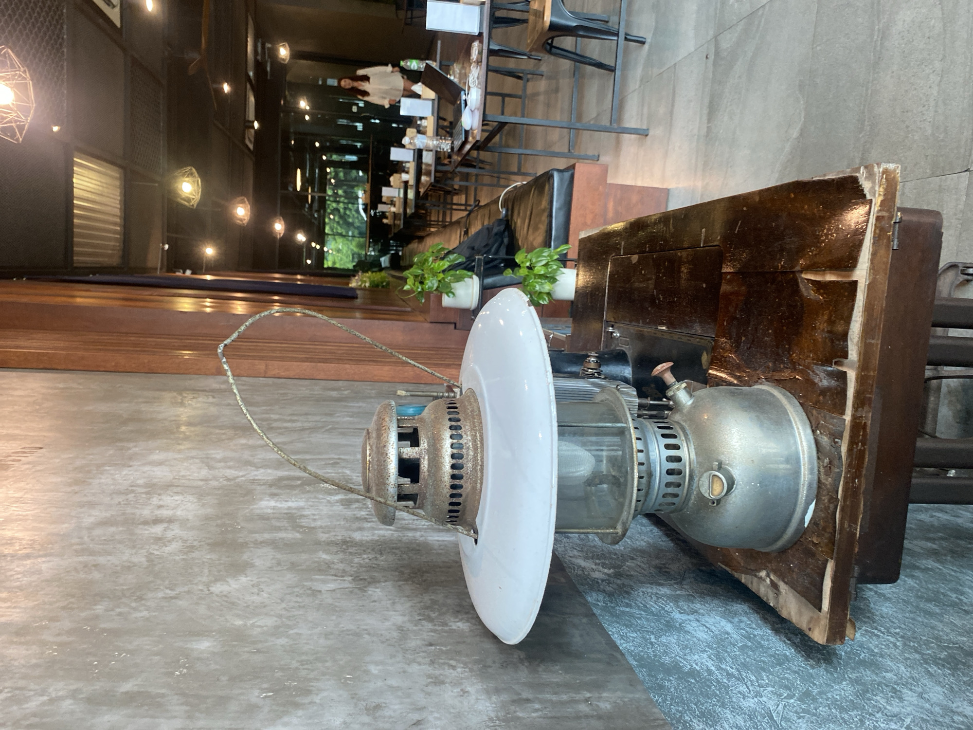 The image shows an antique oil lamp on a gray surface, possibly a floor. The lamp has a white circular shade and a tarnished metal body. It is placed next to a weathered, dark wood structure, which may be part of a table or cabinet.
In the background, a partially visible interior features a row of tables with people sitting at them, suggesting a cafe or restaurant setting. The decor includes hanging lights and indoor plants. Through the windows, there is a glimpse of greenery, indicative of an outdoor area or foliage outside the building.
The location is Phuket City Municipality, Thailand. The presence of furniture, lighting, and people suggests the image was taken in a public or commercial establishment in the city.