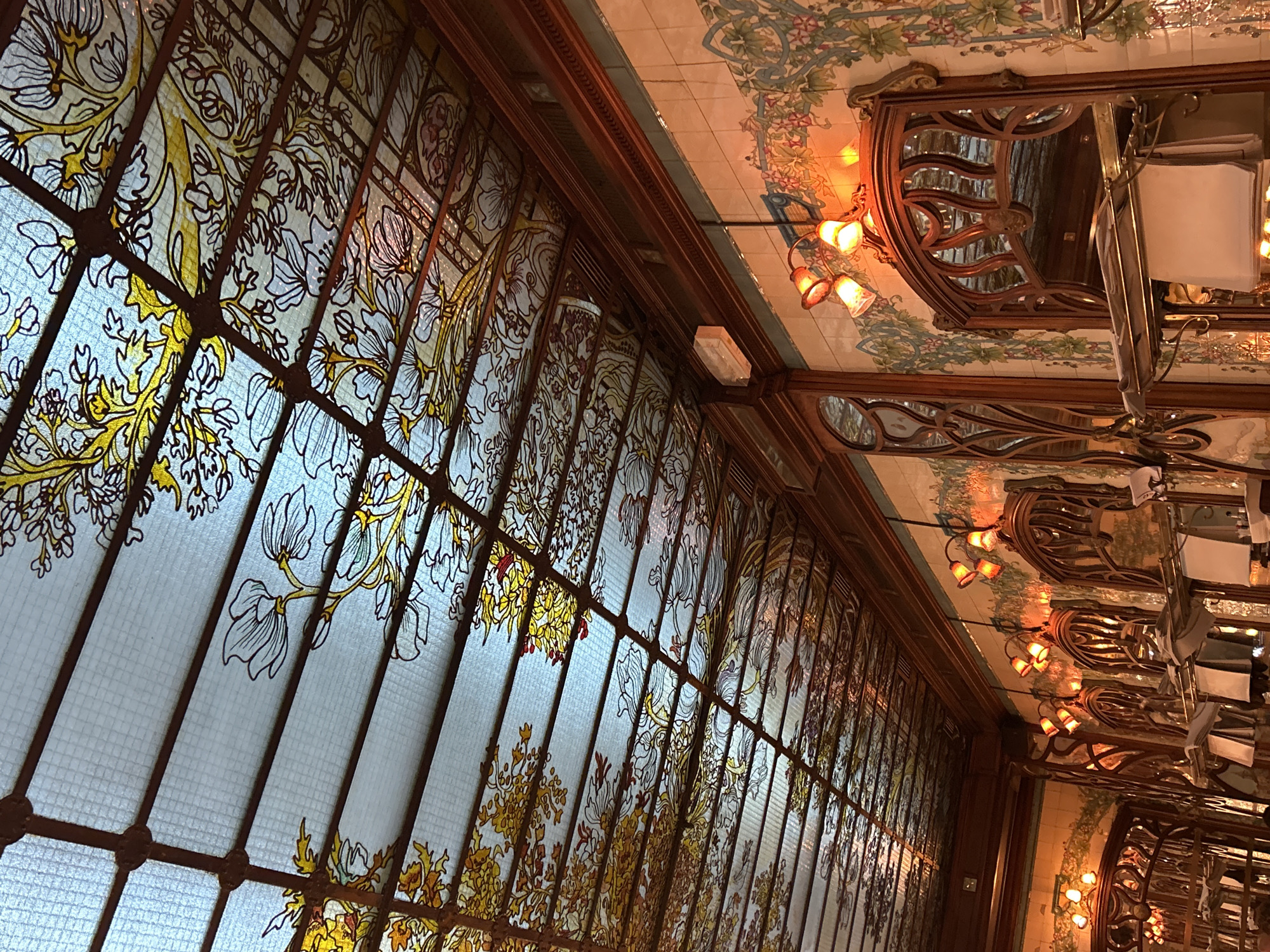 The image depicts the interior of a Parisian restaurant, likely Maxim's or a similar establishment known for its Art Nouveau design. The focus is on a large stained-glass window decorated with floral motifs in yellow and white against a blueish background, divided by dark, ornate metal frames. The adjacent wall and ceiling feature decorative tiling with floral patterns. A series of hanging lamps cast a warm, orange glow. Tables are visible with white tablecloths and chairs with mirrored backs are reflected on the ceiling. The orientation of the image suggests a perspective looking upwards and along the length of the restaurant. The scene is devoid of people.
