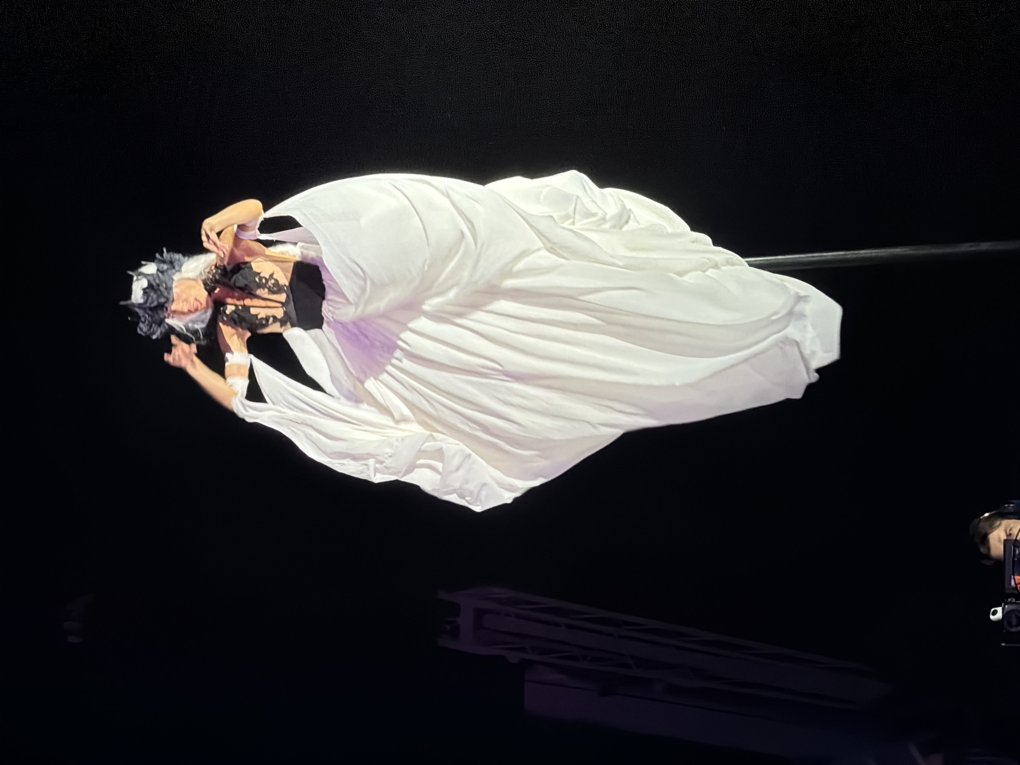 An aerial performer, adorned in a white flowing gown and a black and white headpiece, is suspended mid-air against a dark background. The performer's hands are raised, appearing to adjust the headdress. The voluminous gown billows around the performer, creating a dramatic silhouette. In the lower left quadrant of the image, a person wearing headphones is visible in front of a camera setup, suggesting this may be a staged performance or event. A tall, metallic structure, possibly scaffolding or stage equipment, is also present on the left side of the frame. The overall scene depicts an artistic performance with elements of stagecraft and technical equipment. The location in Odesa, Ukraine, is not discernible from the visual information provided in the image.