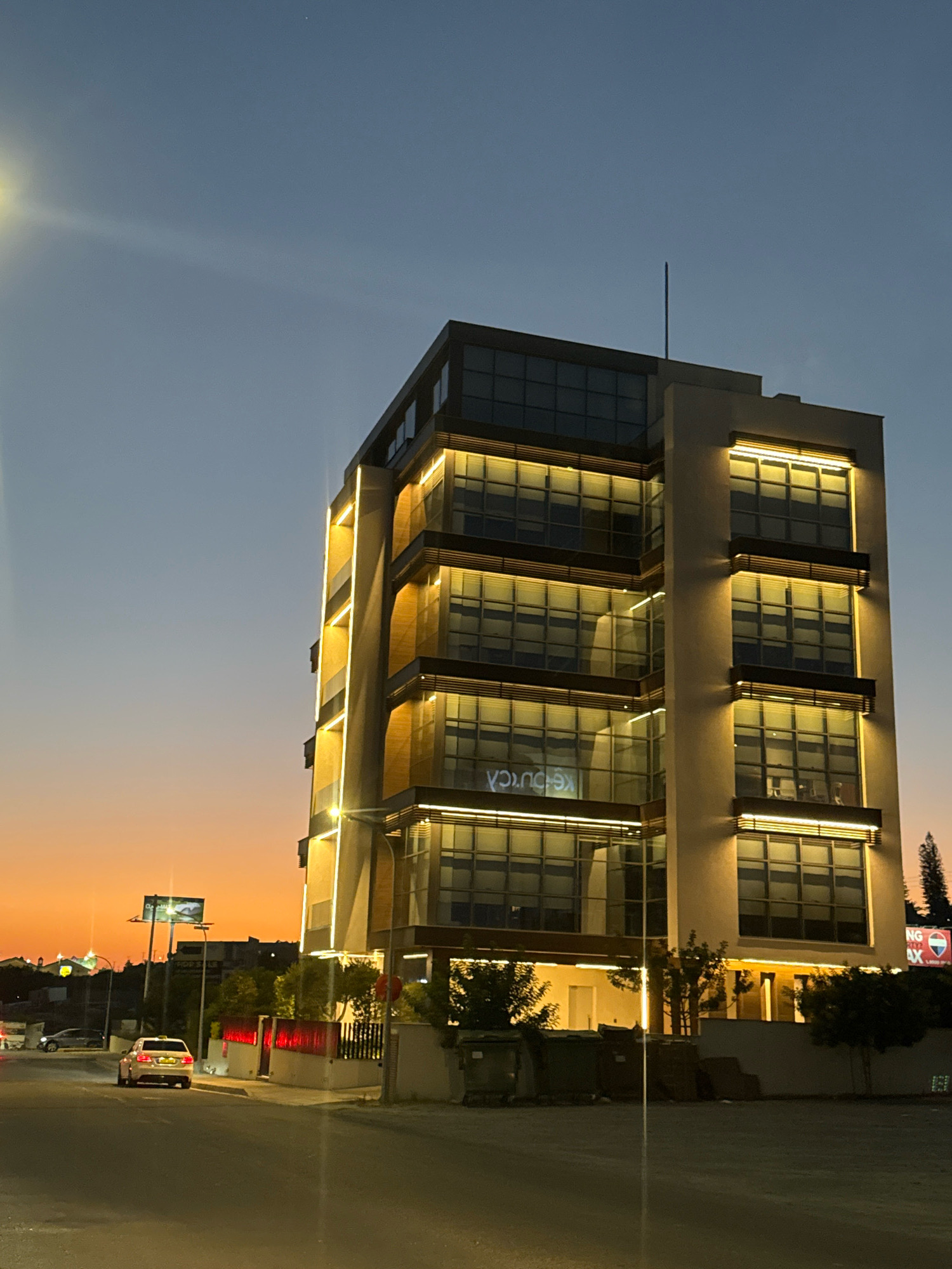 The image shows a modern, multi-story office building at dusk in the Mouttagiaka Community, Cyprus. The building features large windows with a reflective surface, and is illuminated by warm, yellow-toned lights along its facade and at each floor level. The sky transitions from a clear, pale blue at the top to a warm orange and yellow gradient near the horizon, indicative of sunset or twilight. In the foreground, a street runs in front of the building, with a white sedan visible in the left lane. The street appears to be paved and empty except for the vehicle. To the left of the building, there are some trees and what appears to be a fence with red lighting. In the distance, streetlights and other buildings are visible. The overall scene suggests an urban or suburban setting in Cyprus, with the building being a prominent architectural feature illuminated against the darkening sky.