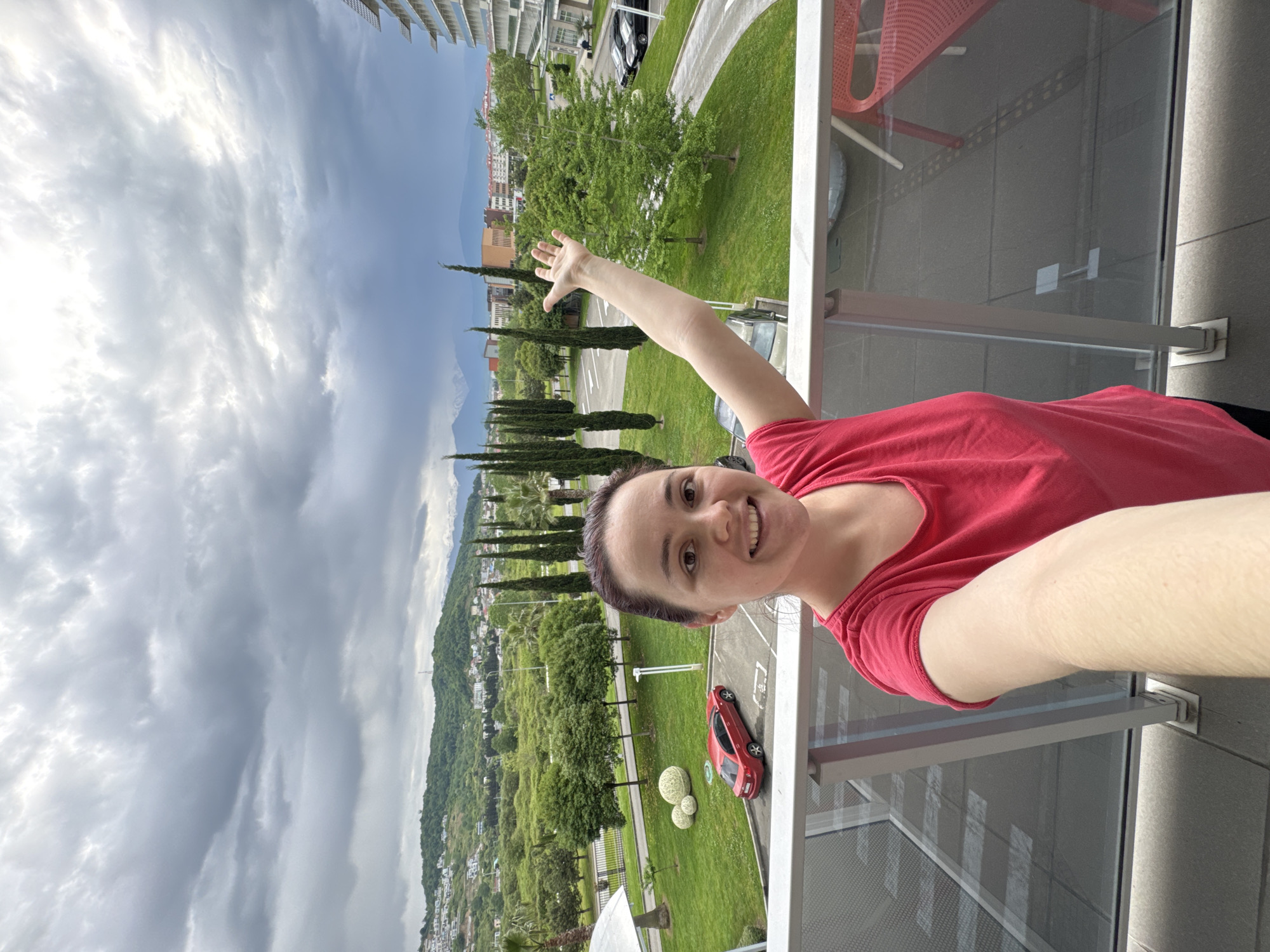 A woman in a red t-shirt is standing on a balcony in Sochi, Russia, waving at the view. The landscape includes tall, narrow trees, green areas, mountains in the distance, and a cloudy sky. A red car and some green spherical objects are visible on the grass. The balcony has a metal railing and glass panels. In the background, there are buildings and infrastructure typical of a city.