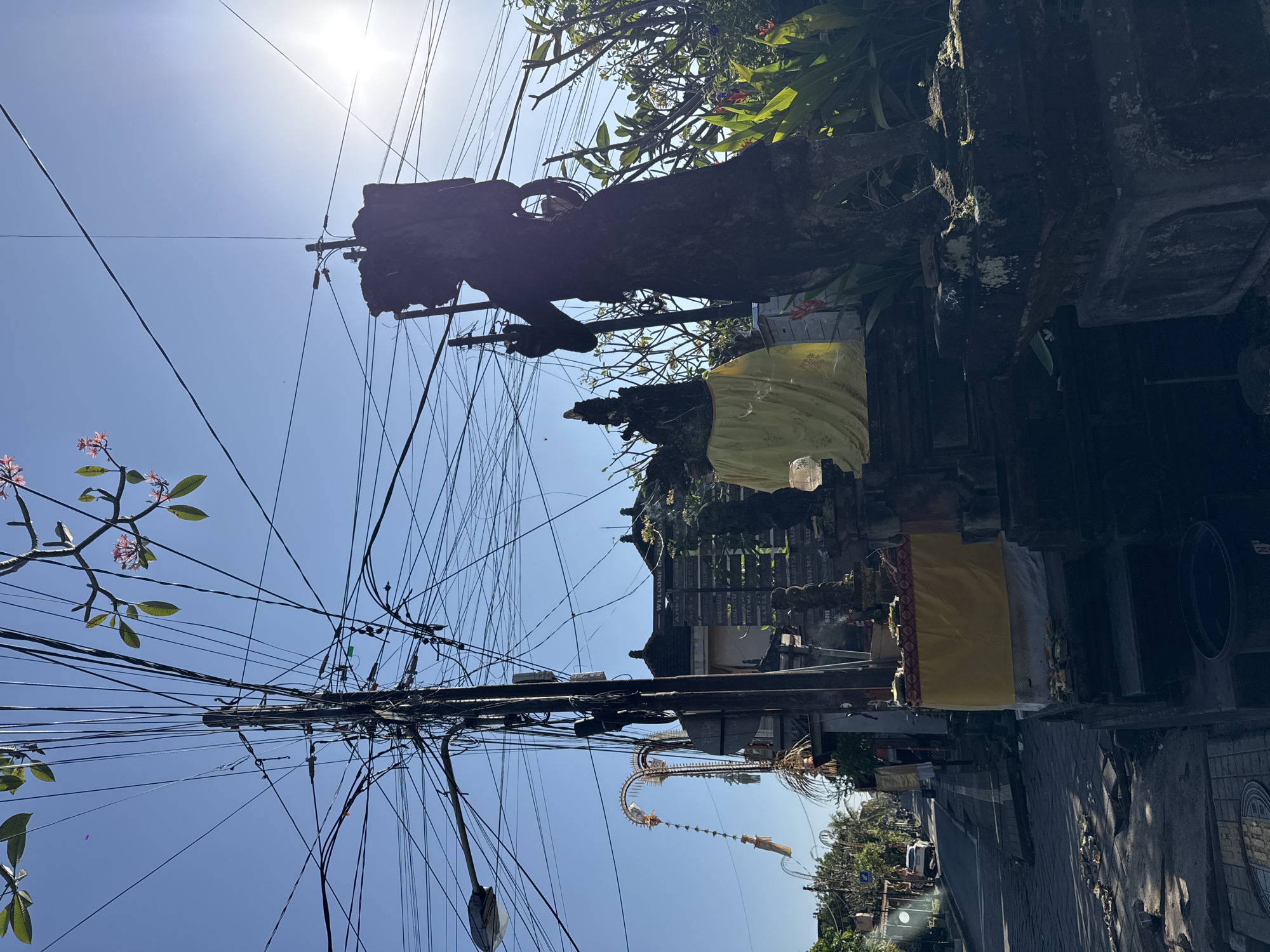 The image captures a sun-drenched street scene in Ubud, Indonesia, likely during the daytime. Dominating the foreground is a large, dark statue, silhouetted against the bright sky, which appears to be a guardian figure with a backpack-like structure on its back and holding a staff. To its left, another statue is draped in yellow fabric, and a tiered stone structure with offerings, including yellow and white cloth, is visible. A paved road with a white line marking the center runs alongside the sidewalk. Further down the road, a white car is parked, and various buildings with traditional architecture are present. The scene is characterized by a complex network of overhead electrical wires, a prominent utility pole, and a street lamp. Lush green foliage and frangipani trees with pink blossoms add a natural element to the urban landscape. The sky is clear and blue, with the sun casting a bright glare.