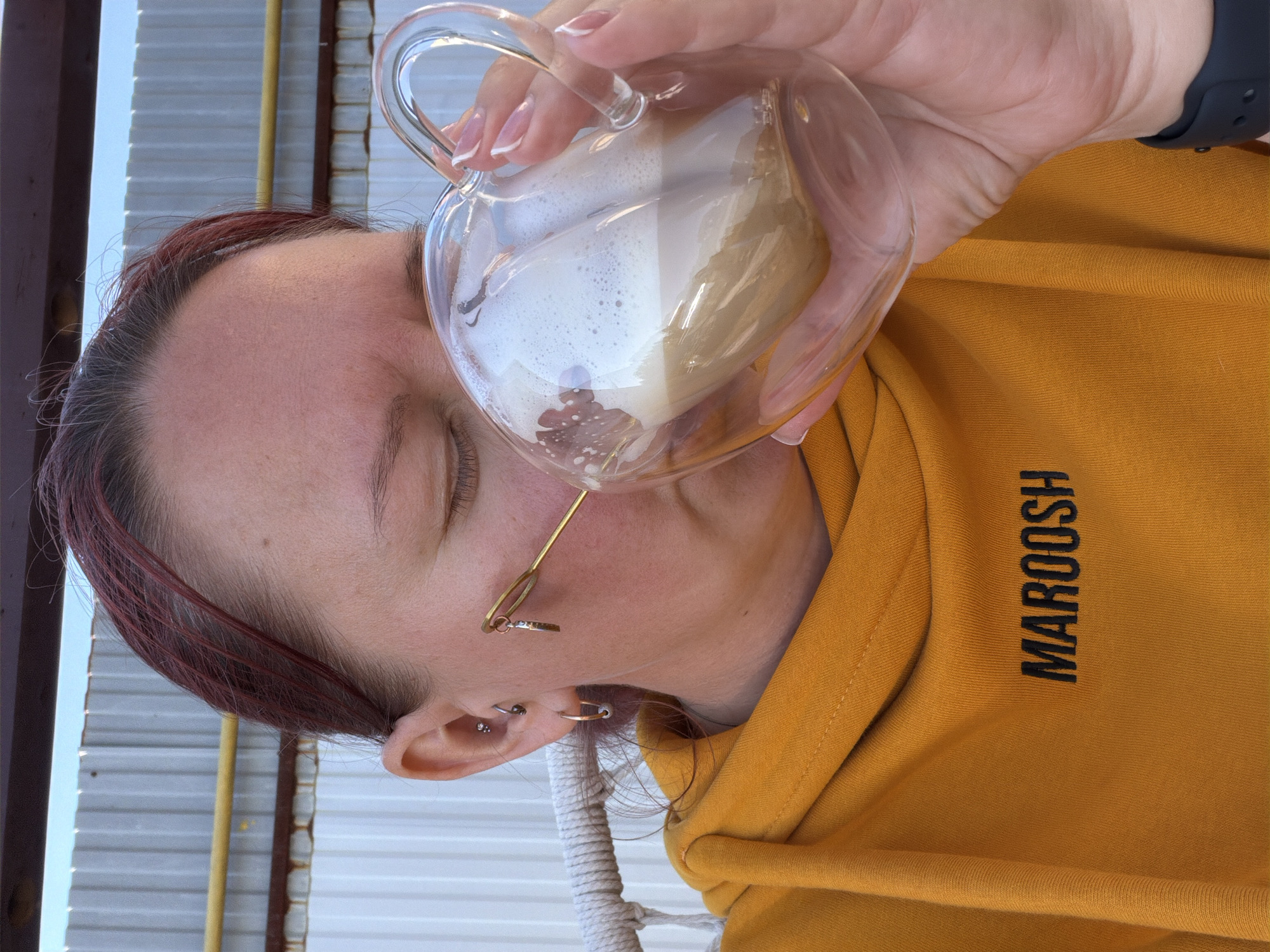 A woman in a mustard-colored hoodie with the word "MAROOSH" vertically printed on it is holding a heart-shaped glass mug filled with a beverage that appears to be a latte. The drink has a foamy layer on top, and the mug is held up close to her face. She has closed eyes, and glasses on. Her left ear has multiple piercings. The background includes what appears to be a building with white siding and a metal frame. The location information mentions the city of Chaikovsky, Russia, but there are no specific landmarks in the image to confirm it.