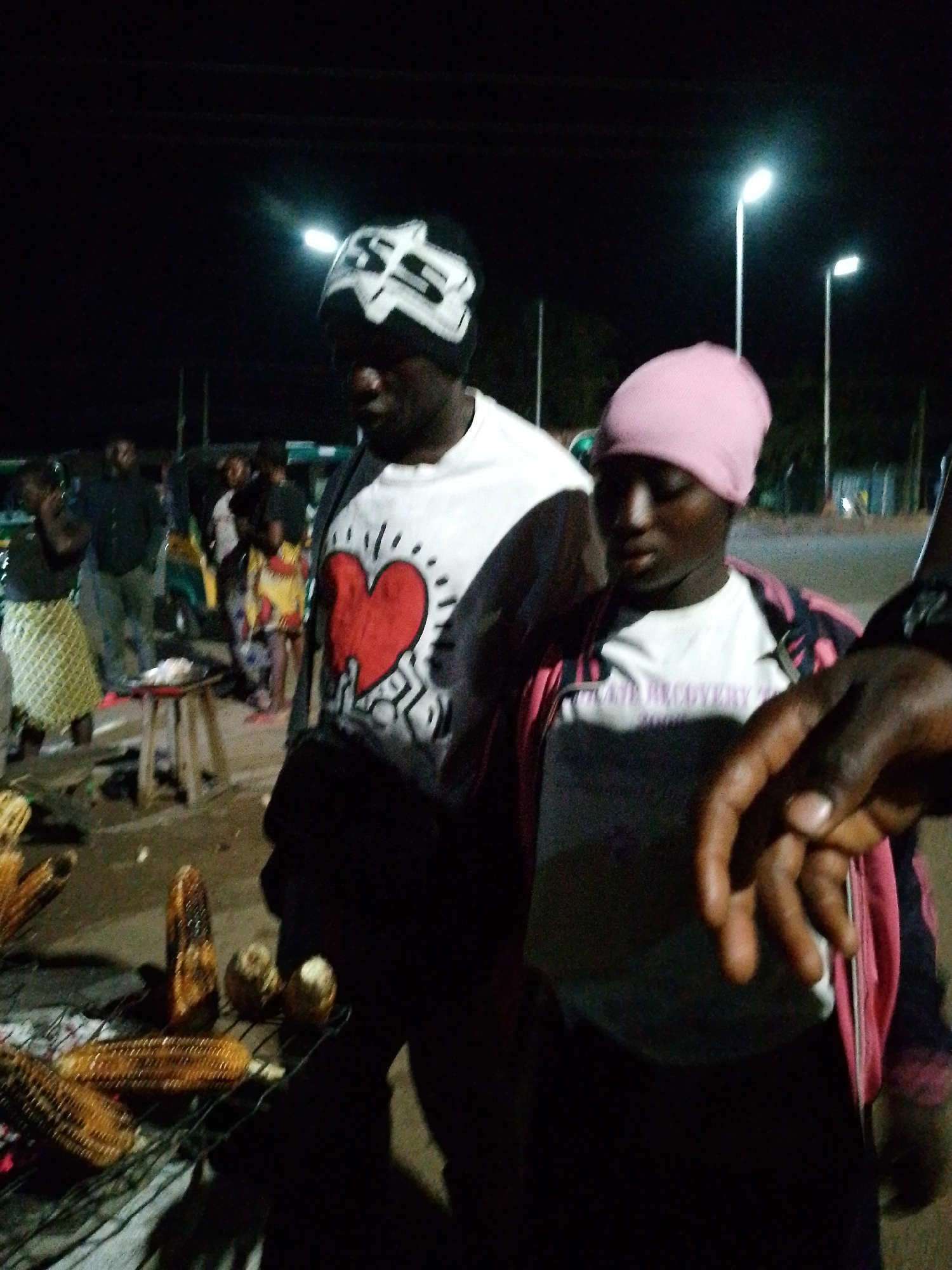 At night in Bukuru, Nigeria, two individuals stand near a grill with roasted corn. The person on the left wears a black beanie with a white pattern and a white t-shirt featuring a red heart graphic. The person on the right is wearing a pink beanie and a pink and black striped jacket over a white t-shirt with faint text. Behind them, other people and a street scene with lampposts are visible. The ground is dirt.