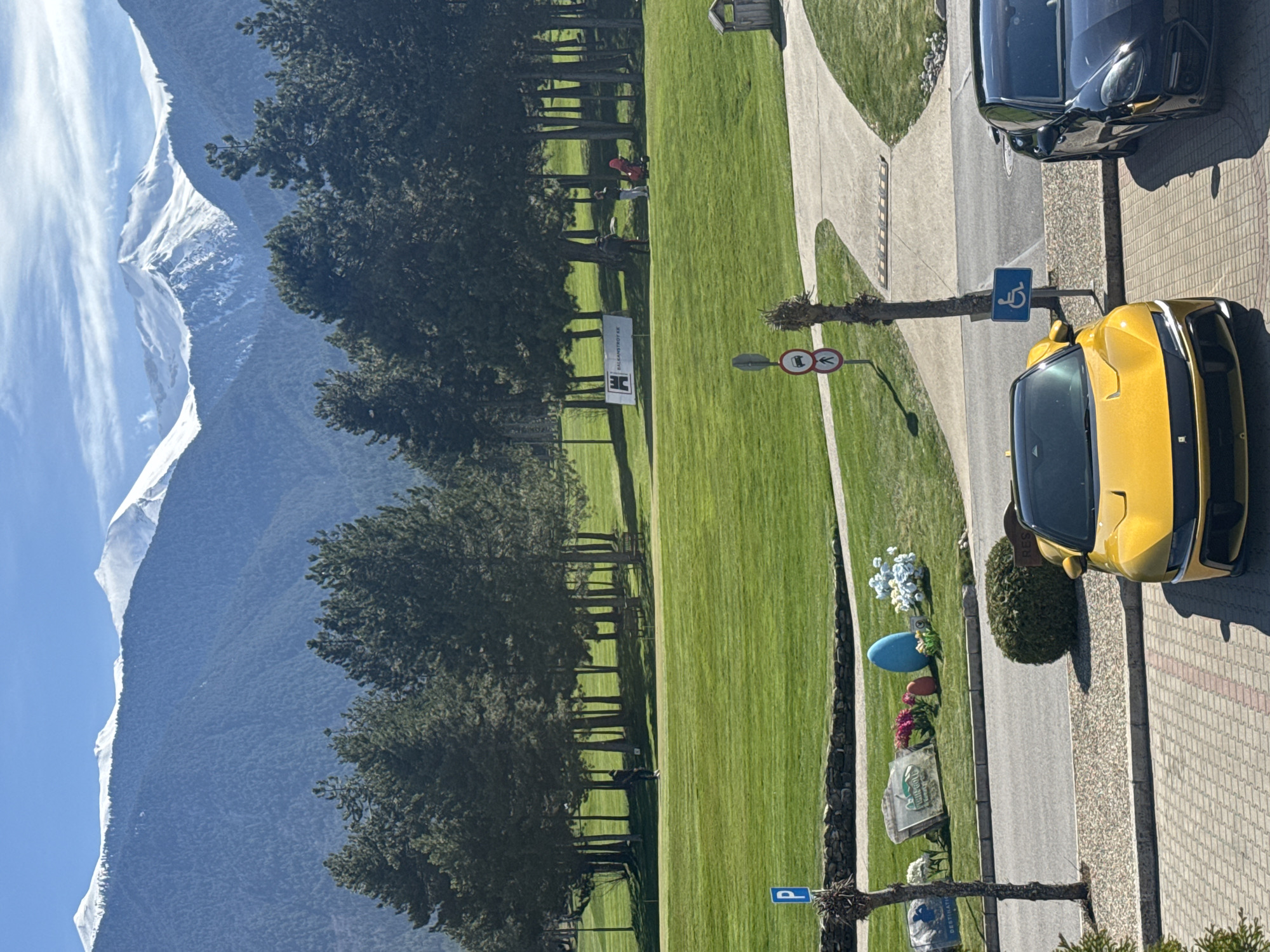 The image shows a view in Razlog, Bulgaria. In the background are mountains covered with snow. Adjacent to the mountains is a row of trees. In front of the trees is a large grassy area. A road runs along the grass with a blue sign with a white wheelchair symbol. There is a yellow car parked adjacent to the sign on a paved area. Also next to the yellow car is a darker car. A pedestrian path with a bench is on the grass. People are walking on the path. The sky is light blue.