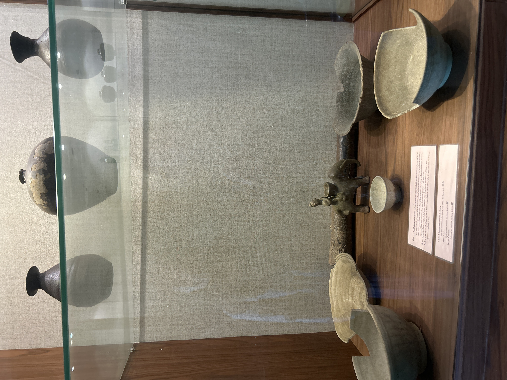 The image shows a museum display case containing various pottery artifacts. On the top shelf, three large ceramic vases are visible, two of which are prominently displayed. The vase on the left is dark grey with a narrow neck, and the vase on the right is a larger, light grey vase with a dark rim and a textured surface. A smaller, dark-colored vase is positioned between them. Below this, on the lower shelf, several more artifacts are arranged.
In the center of the lower shelf, a small, dark bronze figurine of a person riding an elephant is displayed. To the left of the figurine is a large, shallow, pale green ceramic bowl with a wide rim. To the right of the figurine, there are two more ceramic bowls. One is a small, pale green bowl, and next to it is a larger, dark grey, irregularly shaped bowl with a cracked surface. In the foreground, two more pale green ceramic bowls are visible, one on the left with a broken rim and another on the right with a wide, shallow shape.
In front of the pottery on the lower shelf, two small, white display cards are placed. The card on the left contains Thai script and the card on the right has English text. The English text identifies the items as "Layer 1 Sukhothai period pottery B.E. 21-22," "Layer 2 jar," and "Layer 3 Bottle."
The display case has wooden framing on the left and right sides. The background wall of the display case is a textured, light beige fabric. The floor of the display case is a polished wooden surface. The museum appears to be an indoor space, as indicated by the controlled lighting and the presence of a display case. The general setting suggests an archaeological or historical exhibit. The location of the display, Thai Ko Subdistrict, Thailand, places this exhibition within a specific cultural context, likely showcasing local or regional historical artifacts.