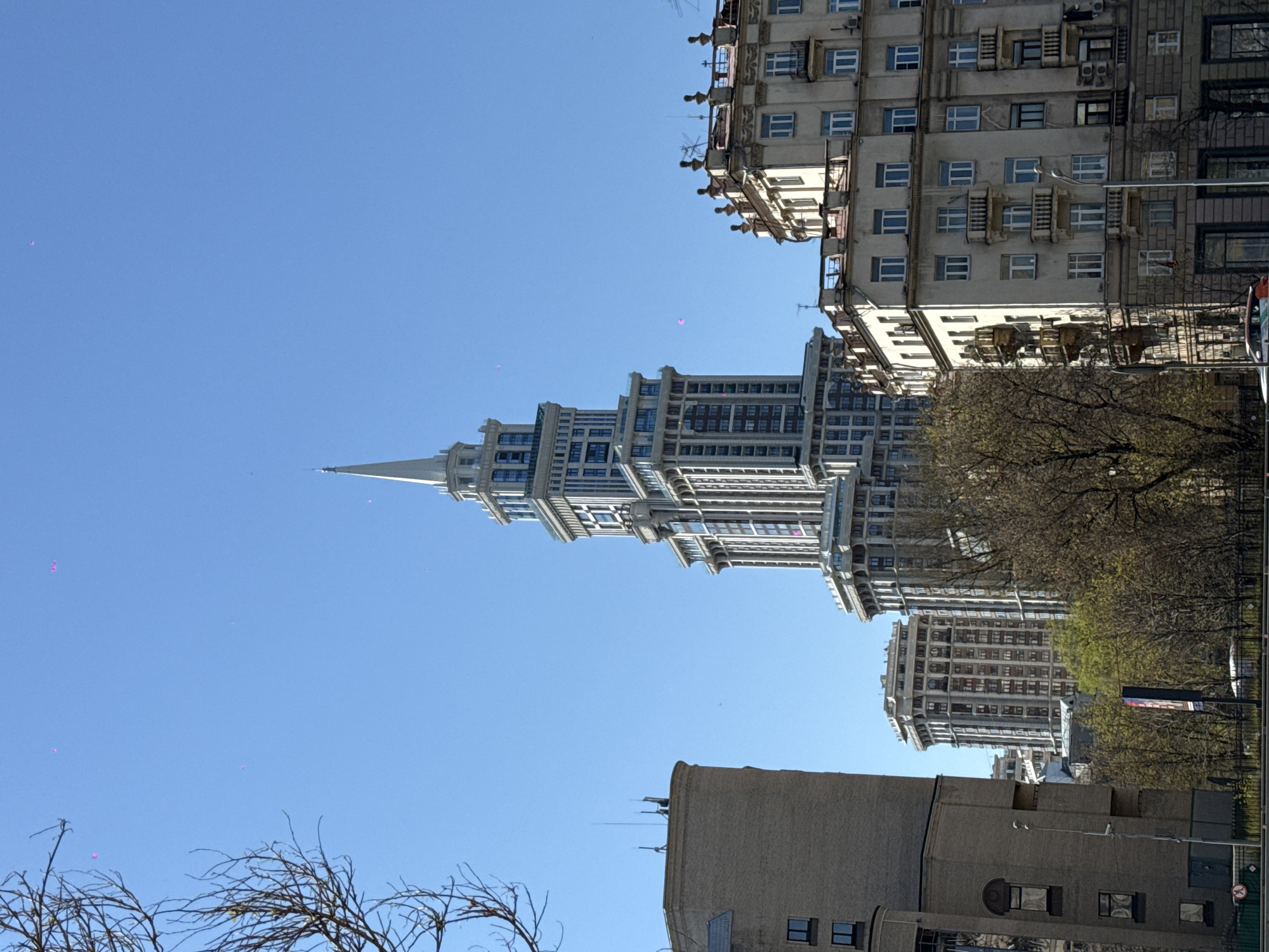 The image depicts a cityscape in Khimki, Russia. A prominent skyscraper with a pointed spire rises in the center of the frame, its architecture displaying multiple tiers and numerous windows. To the right of the skyscraper, several older, less modern buildings are visible, also with multiple floors and windows, partially obscured by trees with budding leaves. In the lower foreground, a brick building with minimal windows stands to the right of bare tree branches. A clear, blue sky serves as the background.