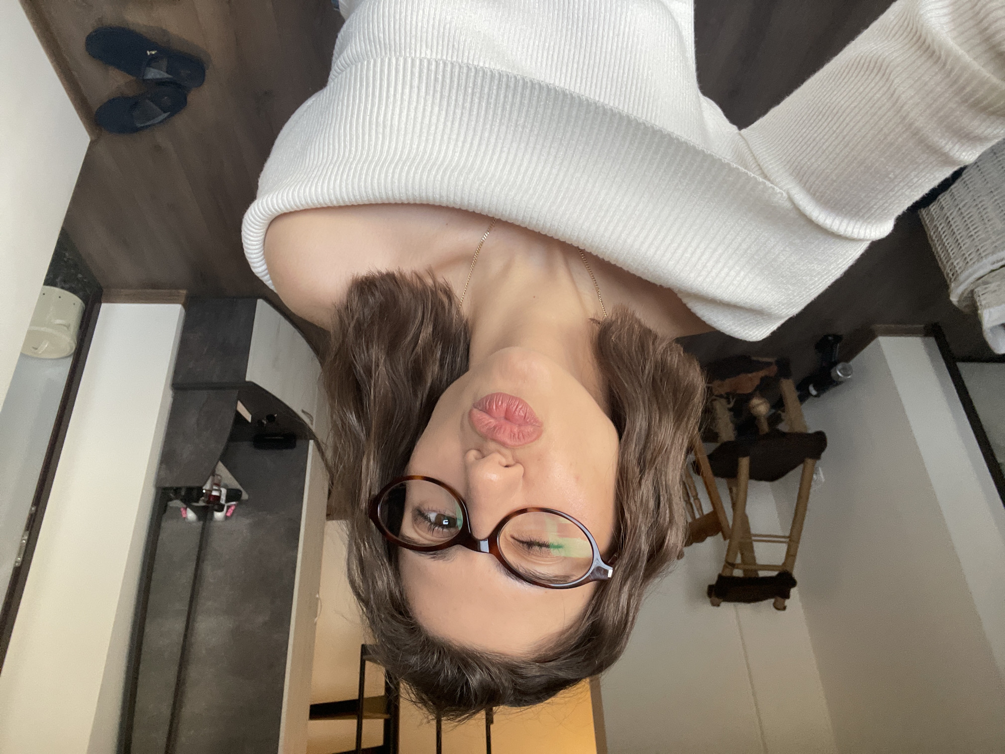 A woman with light brown wavy hair, wearing brown framed glasses and a white ribbed sweater, is taking a selfie indoors. Her lips are pursed in a kissing gesture. She is also wearing a delicate gold necklace. The background includes a wooden floor with a pair of black sandals, a hallway with black and white flooring, stairs, a dark gray rug, and a white wall. A wooden high chair is visible against a white wall. The location cannot be determined to be in the city of Sofia, Bulgaria based on the image alone.