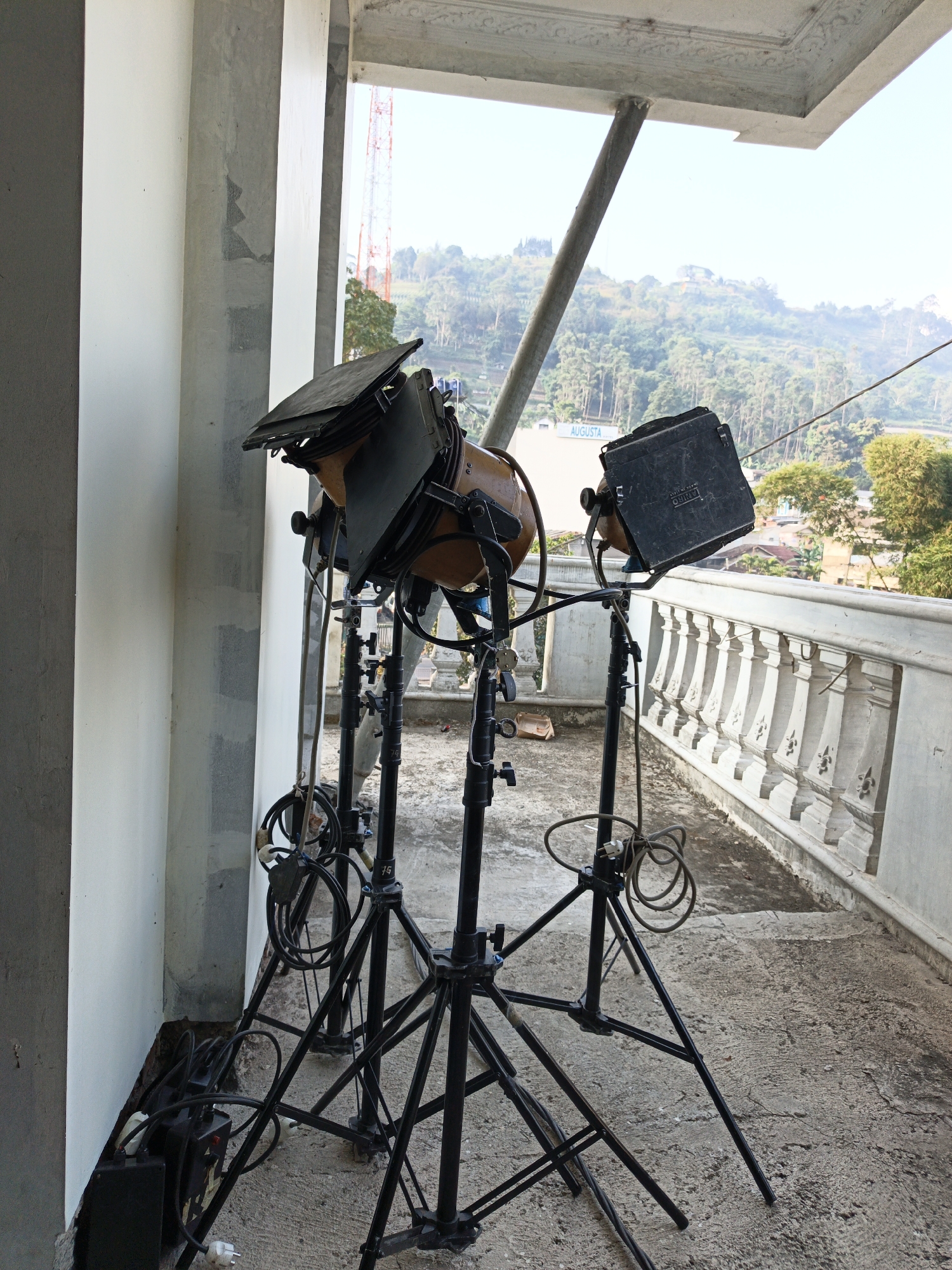 Three professional lighting fixtures, each mounted on a black tripod stand, are positioned on a concrete balcony. The fixtures have adjustable barn doors and appear to be set up for outdoor use. One fixture is a larger, older model with a tan casing and black barn doors. The other two are smaller, black units with similar barn door attachments. Cables are coiled and attached to the tripods, leading to power boxes situated on the balcony floor near the wall. The balcony overlooks a hillside covered in lush green trees and scattered buildings, with a prominent radio tower visible in the distance. The sky is clear and bright. The architecture of the balcony features a white balustrade with decorative spindles. The scene is located in Cibogo, Indonesia, as indicated by the "AUGUSTA" signage visible on a building in the background.