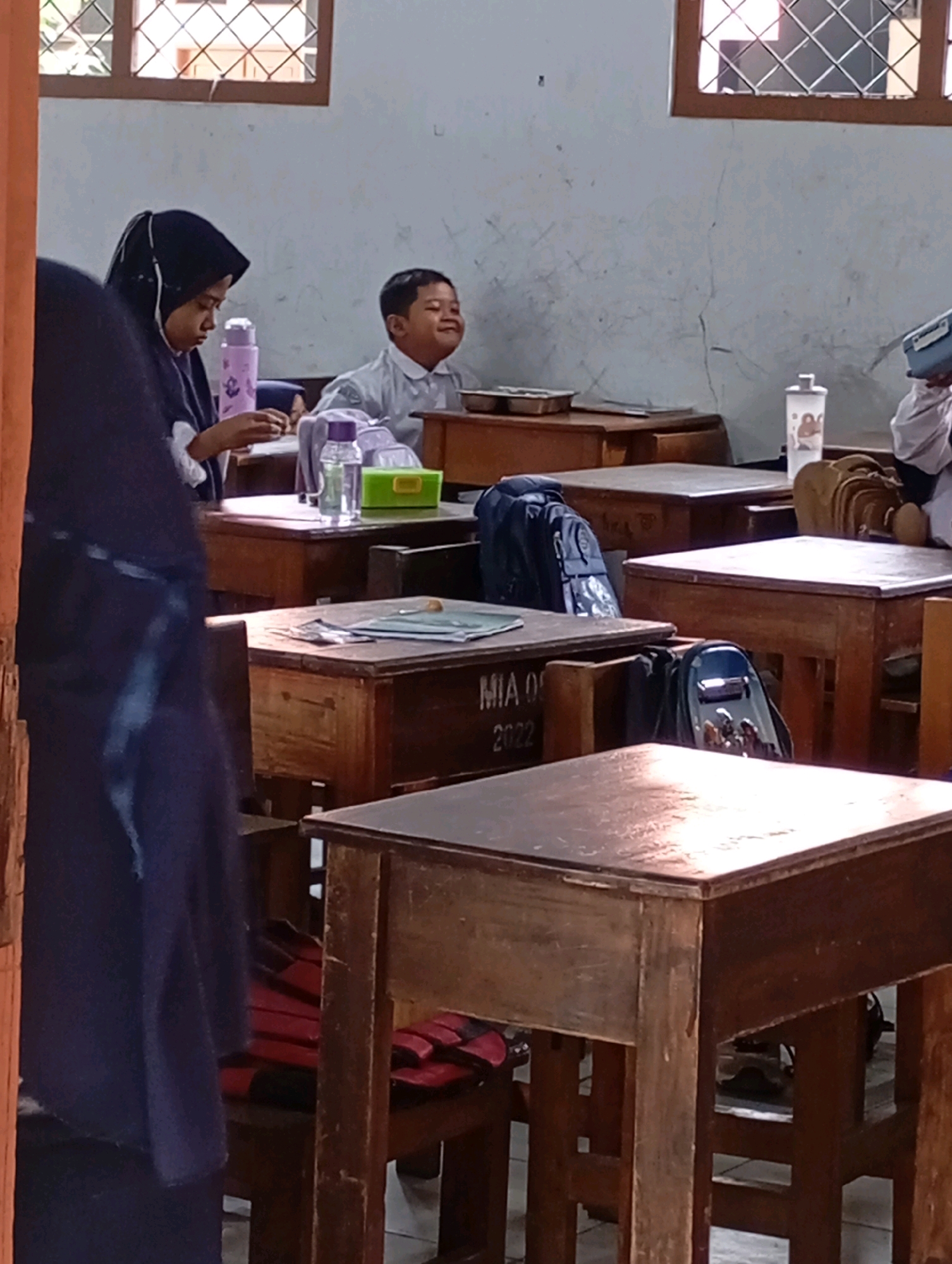 This is a medium-shot, eye-level, indoor photograph taken in a classroom in Babelan, Indonesia. The classroom appears to be old, with wooden desks and a slightly worn chalkboard. Sunlight streams in through a window on the left, illuminating dust motes in the air.
In the foreground on the left, a person wearing a dark blue hijab and a dark blue robe is partially visible, turned away from the camera. They are seated at a wooden desk, with their hands near some papers. To their right, a young boy in a light blue collared shirt is seated at another desk, smiling broadly and looking to the right, perhaps at something or someone outside the frame. A purple water bottle and a green box are on his desk.
Behind the boy, a backpack is visible on his desk. Further to the right, another wooden desk has a backpack and some papers on it. The word "MIA 08" and the year "2022" are written on the side of one of the desks. Other desks and students are visible in the background, as well as a white wall and a window with a diamond-patterned grate. The overall impression is of a typical school day in Indonesia.