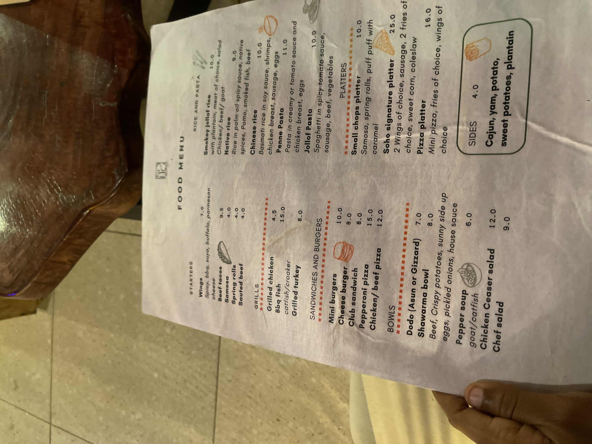 The image shows a menu from a restaurant. The menu is titled "FOOD MENU" and includes sections for "STARTERS", "GRILLS", "SANDWICHES AND BURGERS", "BOWLS", "RICE AND PASTA", "PLATTERS", and "SIDES". The menu items are listed with their prices. The menu is printed on white paper and is somewhat crumpled. The background appears to be a textured surface, possibly a table or a chair. The image is taken from a top-down perspective, with a person's hand visible in the bottom left corner. There are no explicit indicators of the city of Gura Top, Nigeria within the image content itself.