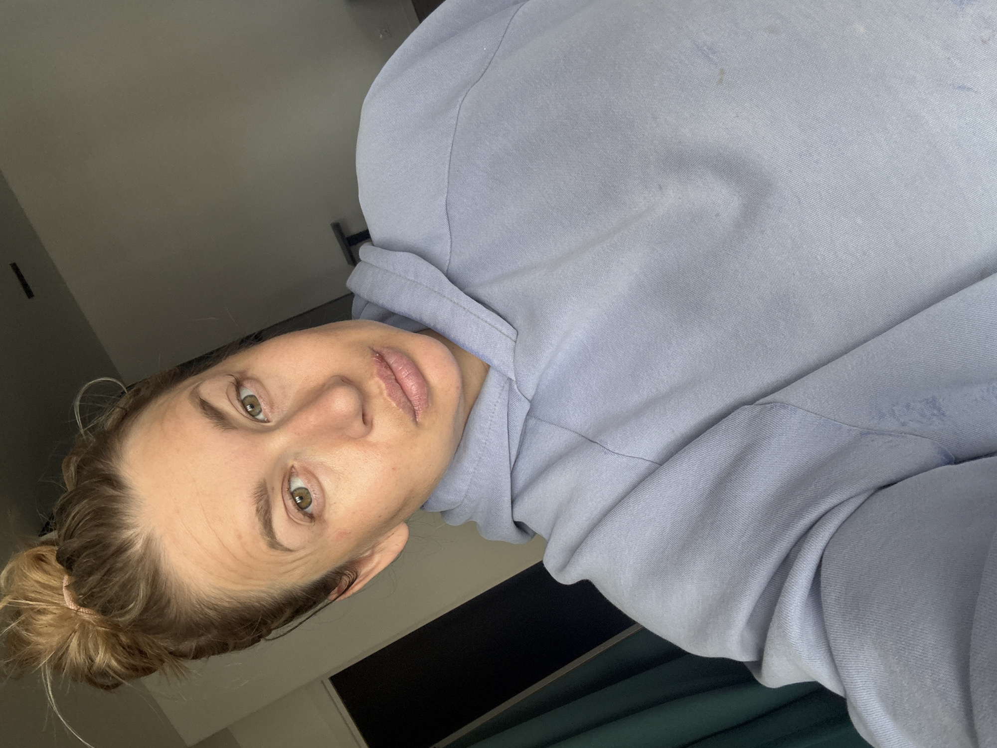 A selfie taken by a light-skinned woman with her hair in a bun, wearing a light blue hoodie. The background appears to be an interior space with a partially visible window and a dark wall. The location is not identifiable as Veliky Novgorod, Russia, based on the provided image.