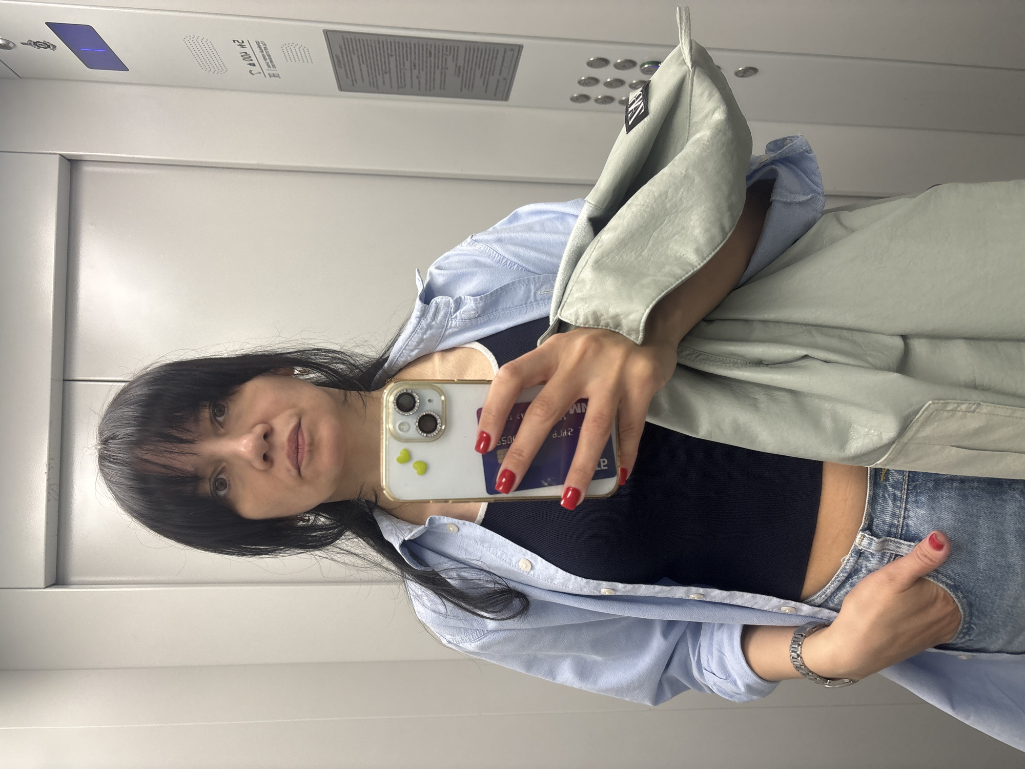 A woman with black hair and bangs is taking a selfie in what appears to be an elevator. She is holding her phone with a gold-colored case and a credit card tucked into the back. She is wearing a black tank top, a light blue button-down shirt, and jeans. She also has on a silver wristwatch. A light green jacket hangs over her arm. The elevator interior is light gray with elevator controls visible in the background.