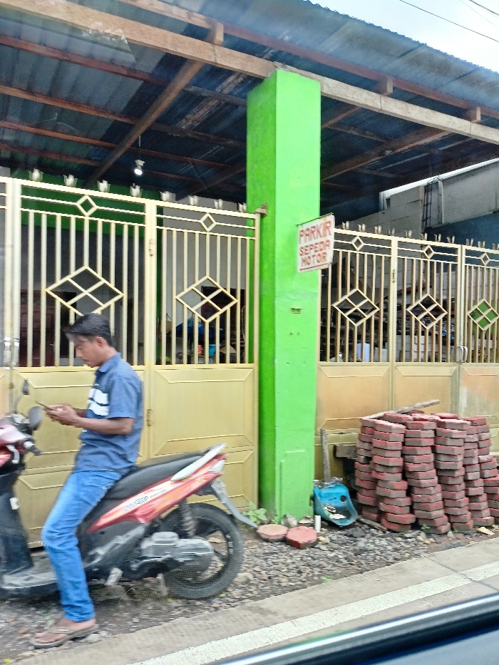 The image captures an outdoor scene, likely roadside, in front of a building. A man, wearing a blue collared shirt, blue jeans, and sandals, is seated on a red and black scooter (moped). He is looking down at a smartphone he holds in his hands, seemingly engrossed in it.

Behind the man is a light gold-colored metal gate with decorative diamond patterns, forming the entrance to a building. Above the gate, there's a roof structure made of corrugated metal sheets supported by wooden beams. A prominent, bright green square pillar stands beside the gate. Attached to this pillar is a white sign with red lettering that reads "PARKIR SEPEDA MOTOR," which translates from Indonesian to "Motorcycle Parking."

To the right of the green pillar and gate, a large stack of red-brown interlocking bricks is neatly piled on the ground, suggesting ongoing construction or landscaping work. A small blue object, possibly a bucket or tool, is visible near the base of the brick pile. The ground in the foreground is rough, appearing to be gravel or dirt next to a paved area. The lighting suggests it is daytime, possibly overcast, as there are no harsh shadows. The overall setting and the Indonesian text on the sign are consistent with the provided location context of Ngoro, Indonesia.