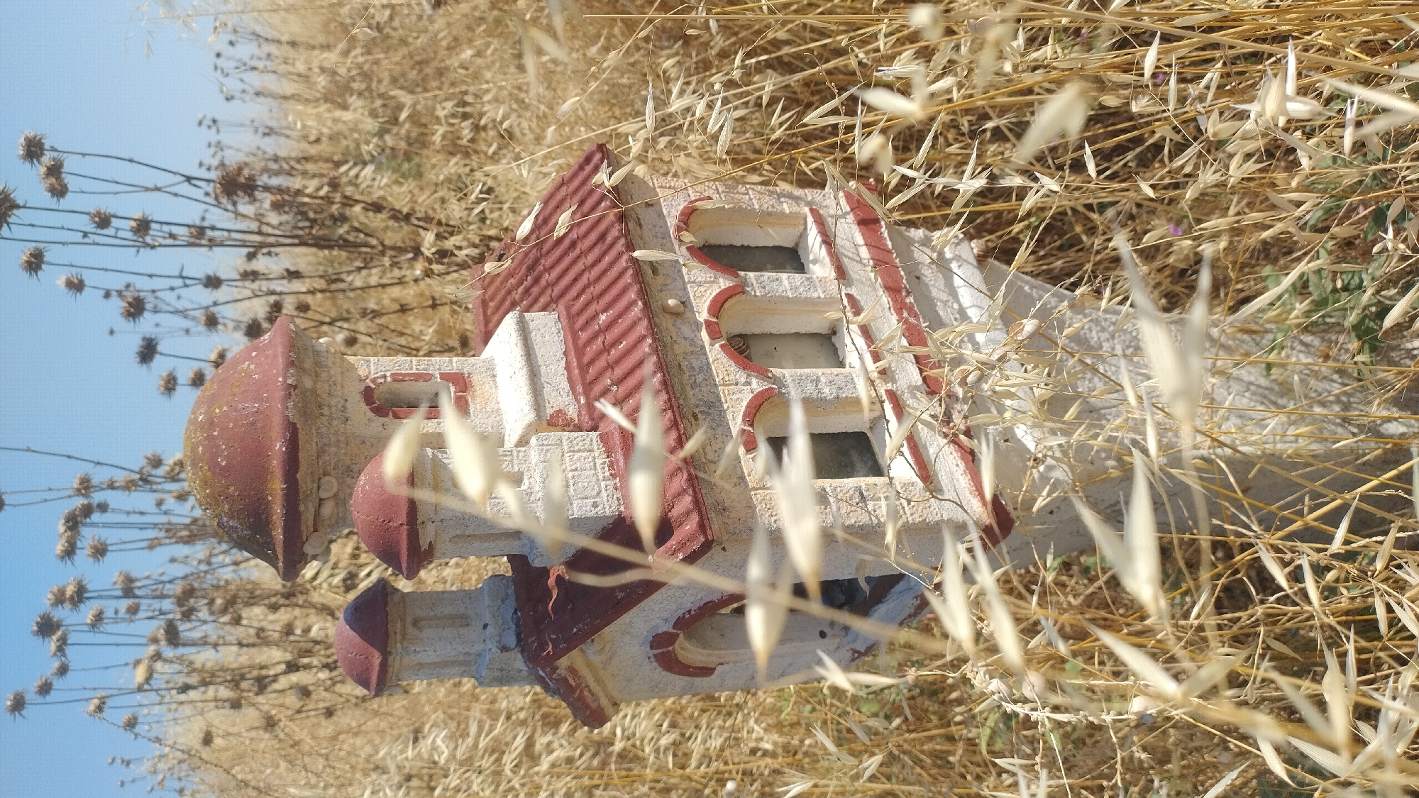 The image displays a weathered miniature church-like structure, positioned amidst dry vegetation under a clear blue sky, located in Thermi, Greece.
The central subject is a small architectural model or shrine, predominantly off-white with reddish-brown accents. It features a large, moss-tinged reddish-brown dome at its highest point, flanked by two smaller, similarly colored domed towers to its left. The main body of the structure is constructed to mimic light-colored stone blocks, with horizontal and vertical trim in a darker red color. Several arched openings, resembling windows or niches, are visible on its front and side. The roof beneath the main dome is reddish-brown and appears to have a textured, possibly tiled, surface. The entire miniature building is elevated on a light-colored, square-sectioned stone pedestal, the base of which is largely obscured by foliage. Small, pale snail shells are noticeable on the surface of the main dome.
The immediate surroundings and midground are densely populated with dry, golden-yellow grasses and weeds, some of which are out of focus in the foreground, creating a shallow depth of field. In the background, taller, dried-out plants with distinct seed heads, possibly thistles or similar species, rise against the sky. The sky itself is a uniform, light blue, devoid of clouds. The scene is illuminated by strong natural light, indicating an outdoor setting, likely under direct sunlight. No people or animals are present.