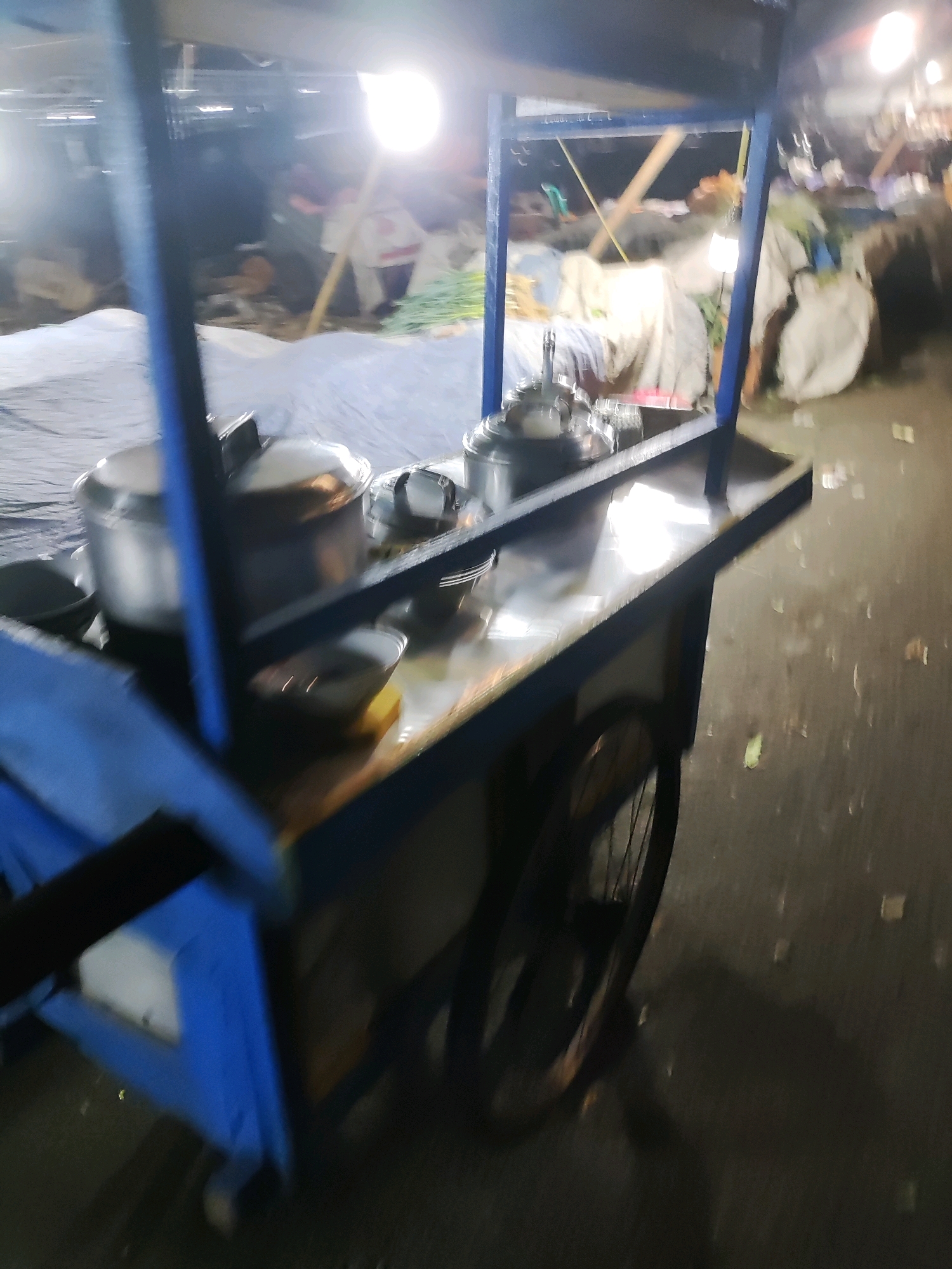 An outdoor nighttime scene in East Jakarta, Indonesia, features a food cart with a blue frame and a metal serving area. The cart is laden with various stainless steel pots and bowls, some containing food or liquids. A large, covered pot sits prominently on the cart. In the background, out-of-focus figures are visible, along with tents and market stalls suggesting a bustling night market or food stall area. Bright overhead lights illuminate the scene, creating reflections on the metal surfaces of the cart. A wheel is attached to the cart, indicating it is mobile. The ground appears to be wet or reflective, and scattered leaves are present.